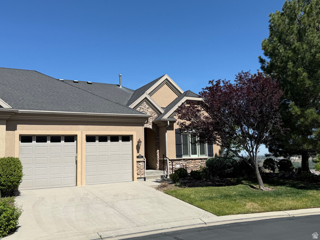 Stucco and stone exterior featuring a two-car garage, dark asphalt shingle roof, and a mature tree providing shade