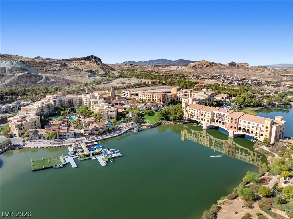 45 Garibaldi Way Henderson, NV 89011 - Photo 66 of 74 A vibrant lake community with a bridge-like building connects various waterfront homes, framed by distant hills.