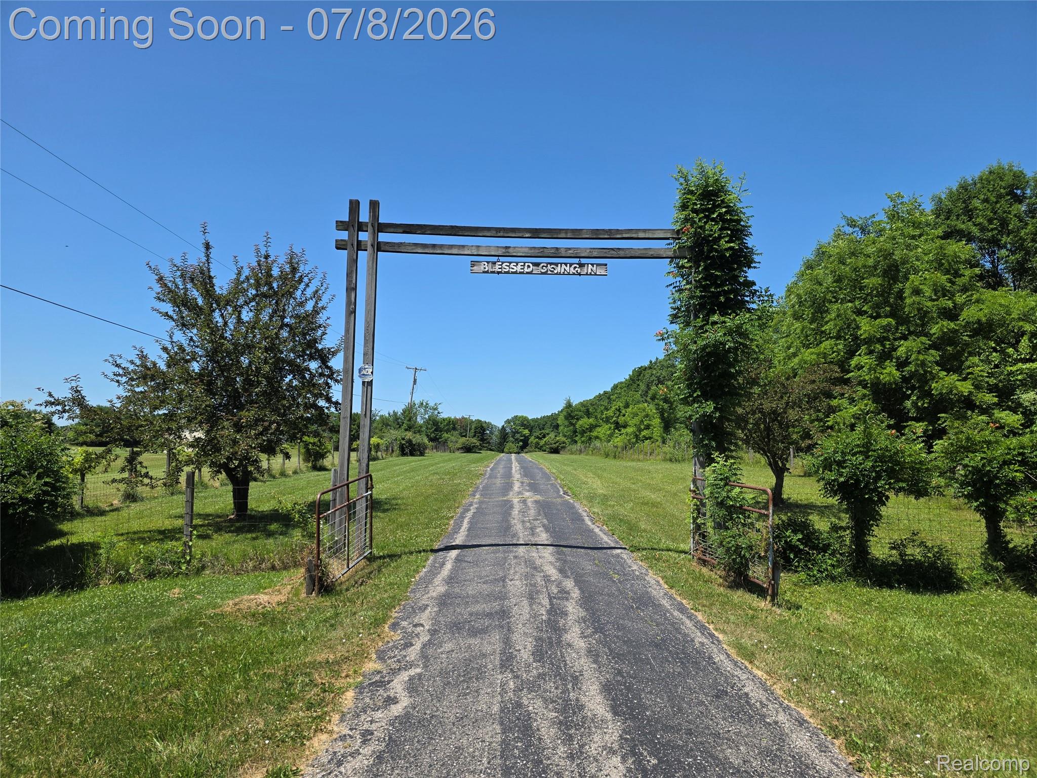 3581 North Burkhart Road Howell, MI 48855 - Photo 2 of 15 Paved driveway bordered by expansive green lawns and mature trees, featuring a rustic wooden entry gate and distant natural landscaping
