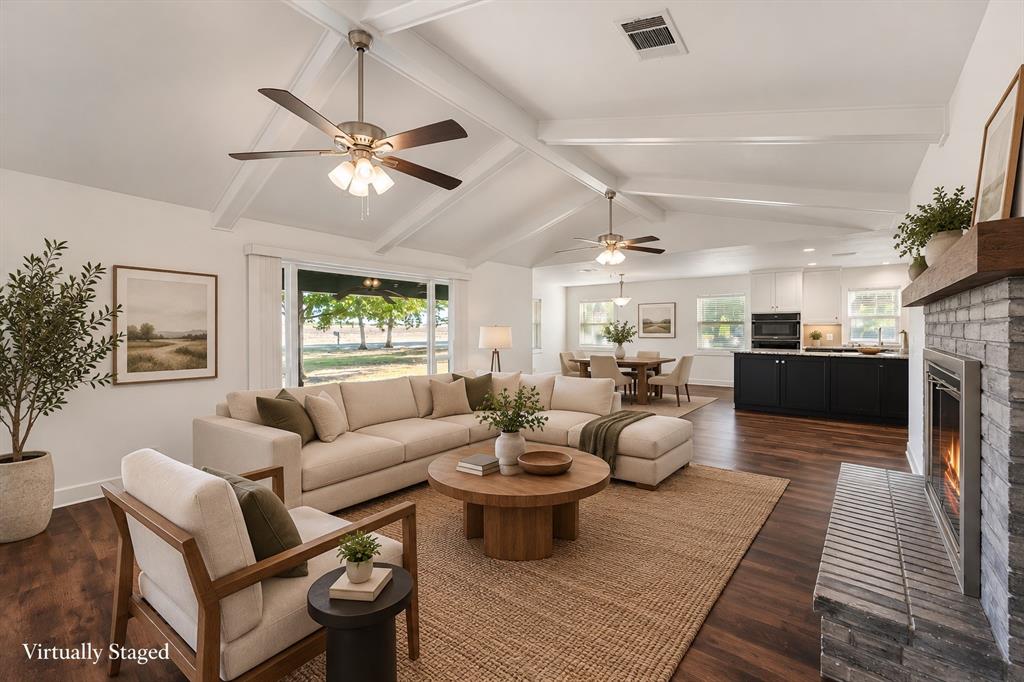 Vaulted ceiling with exposed beams, wood-finish flooring, stone-clad fireplace with wood mantle, built-in kitchen cabinetry, and large sliding glass doors
