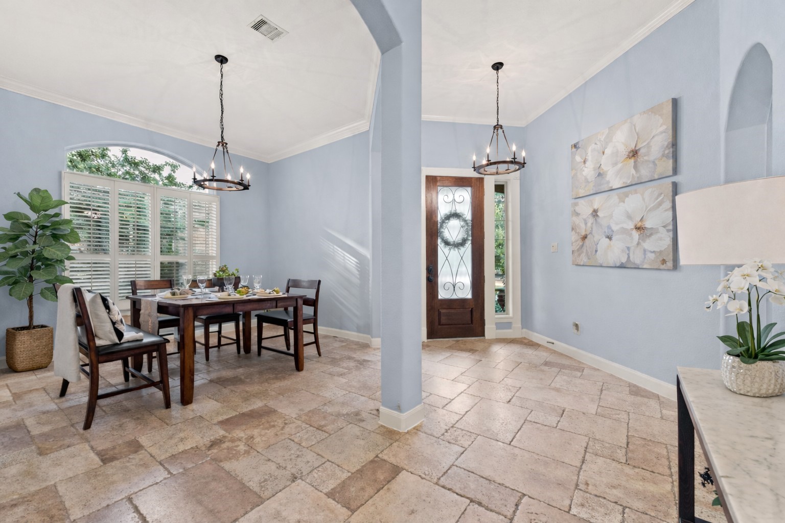 70 Valera Ridge Drive Spring, TX 77389 - Photo 10 of 37 Bright dining and entry area with light blue walls, tile flooring, and two elegant chandeliers. Features a wooden dining table, large windows with shutters, and tasteful floral artwork. Inviting and spacious.
