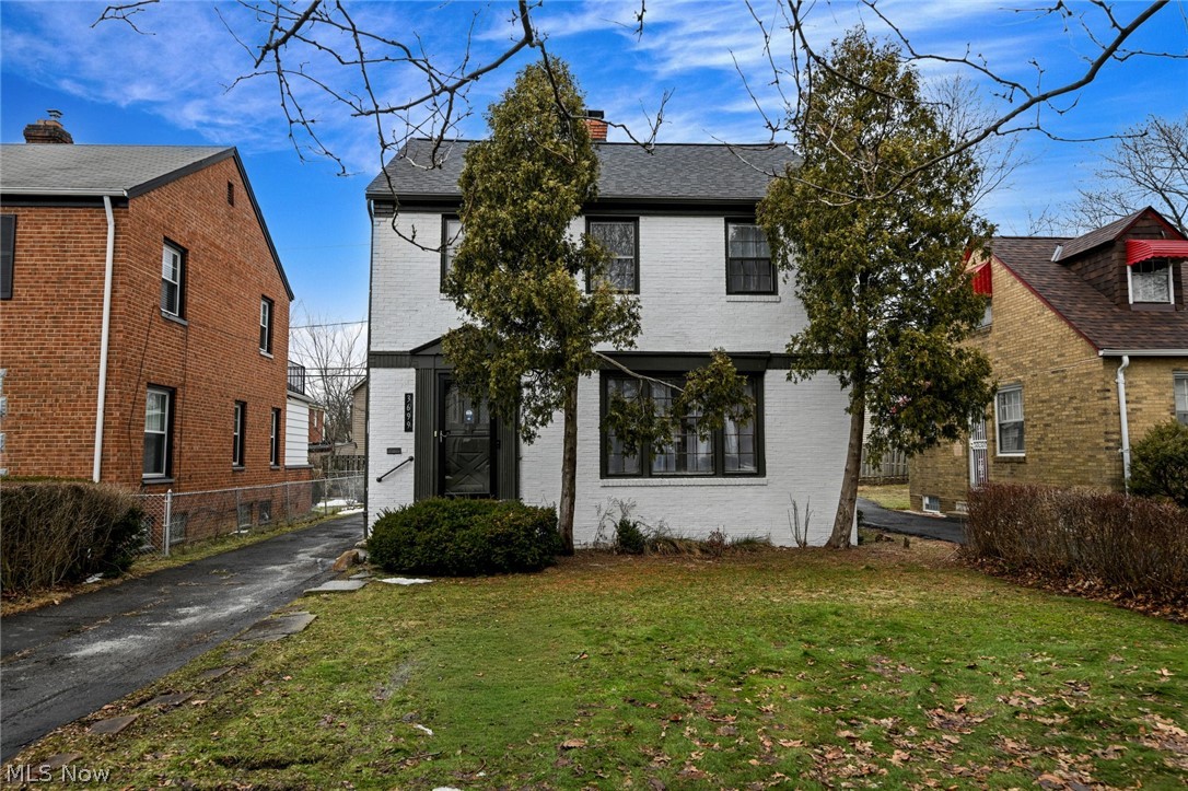White brick exterior with dark trim windows, a front lawn, paved driveway, and mature front trees