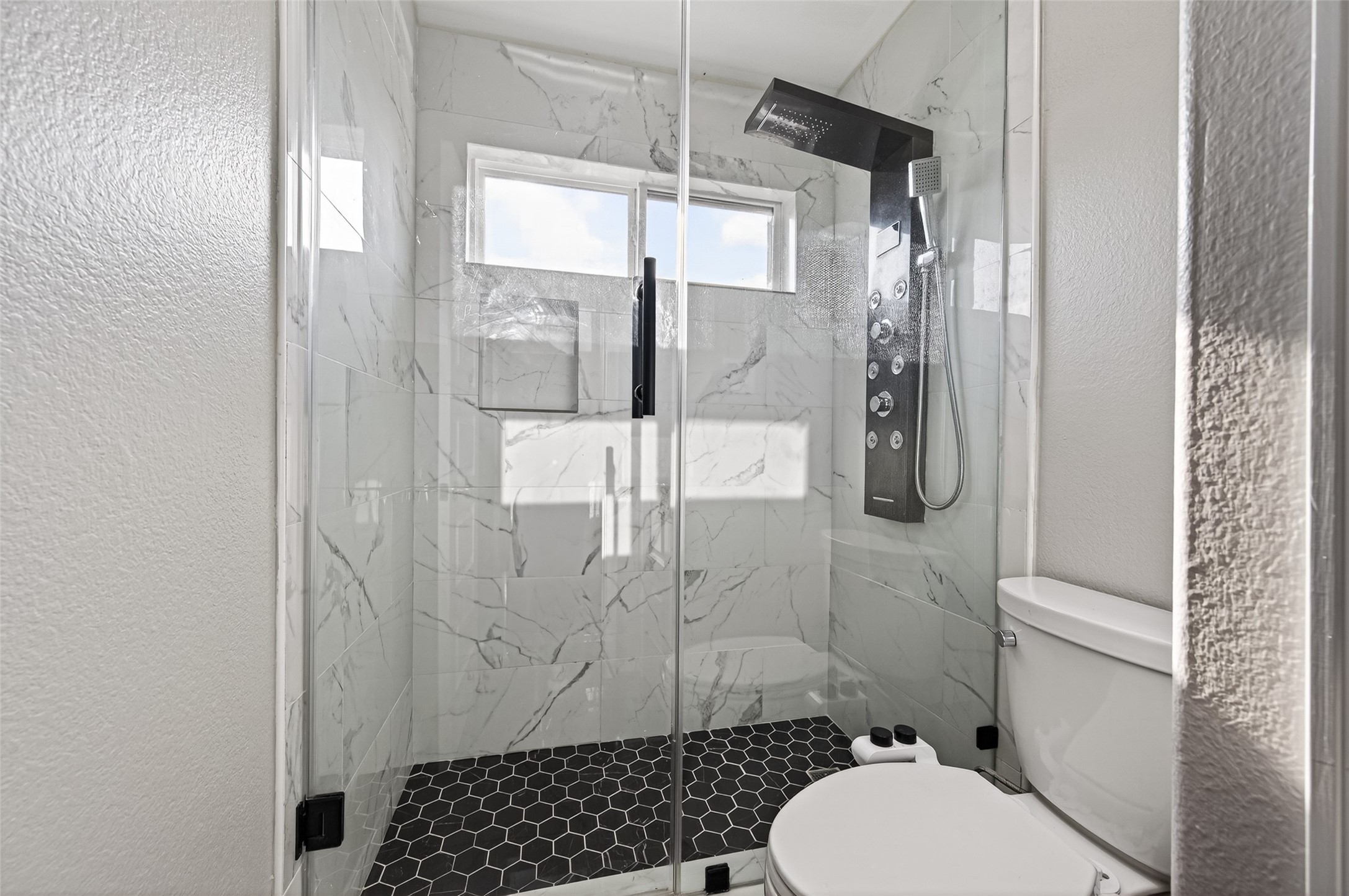 7003 Bristol Memorial Drive Spring, TX 77379 - Photo 33 of 40 Beautifully updated walk-in shower featuring oversized marble-look tile, modern black hexagon flooring, and a sleek glass enclosure for a clean, contemporary finish. The spa-inspired shower panel includes a rainfall head and multiple body jets, creating a luxury experience ideal for busy mornings or relaxing evenings. A transom window adds natural light while maintaining privacy, making this refreshed upstairs bath a standout feature in this spacious Spring TX home.