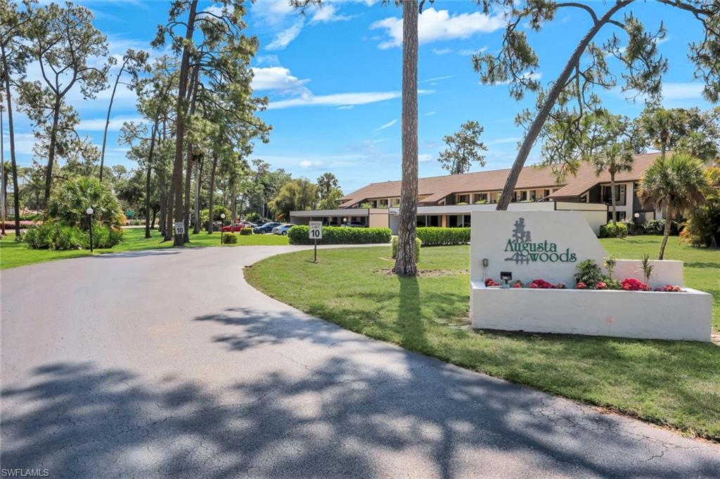 Gated community entrance with signage, mature landscaping, and curved asphalt driveway
