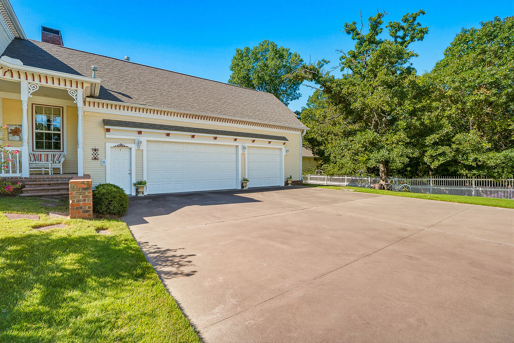 1009 Southeast County Road Ada, OK 74820 - Photo 54 of 92 Expansive driveway, attached multi-car garage, covered porch with decorative trim, brick column accent, and a fenced yard with mature trees