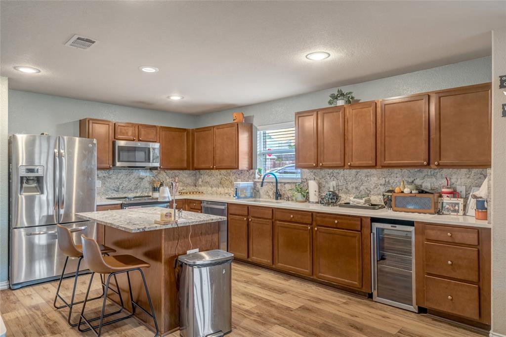 3216 Jetranger Road Fort Worth, TX 76053 - Photo 13 of 35 Kitchen with brown cabinetry, a textured wall, appliances with stainless steel finishes, light wood finished floors, and wine cooler