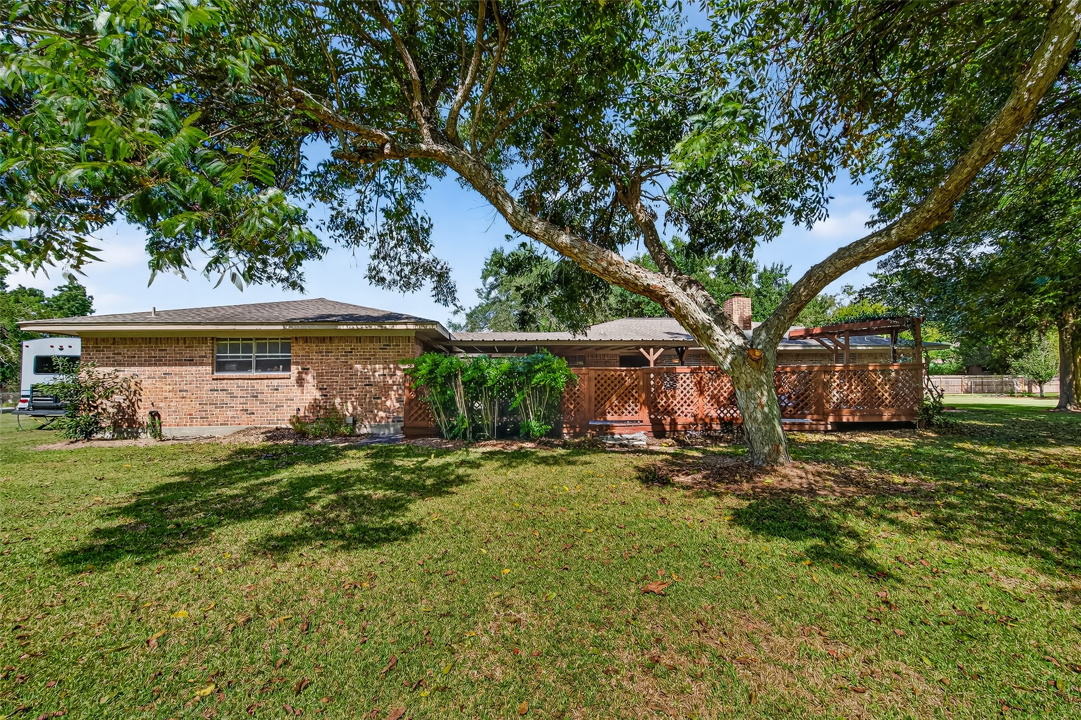 12021 7th 1/2 Street Santa Fe, TX 77510 - Photo 7 of 49 Charming brick home with a spacious backyard featuring lush grass and mature trees. A wooden patio area provides shade and outdoor living space, perfect for relaxation and entertaining.