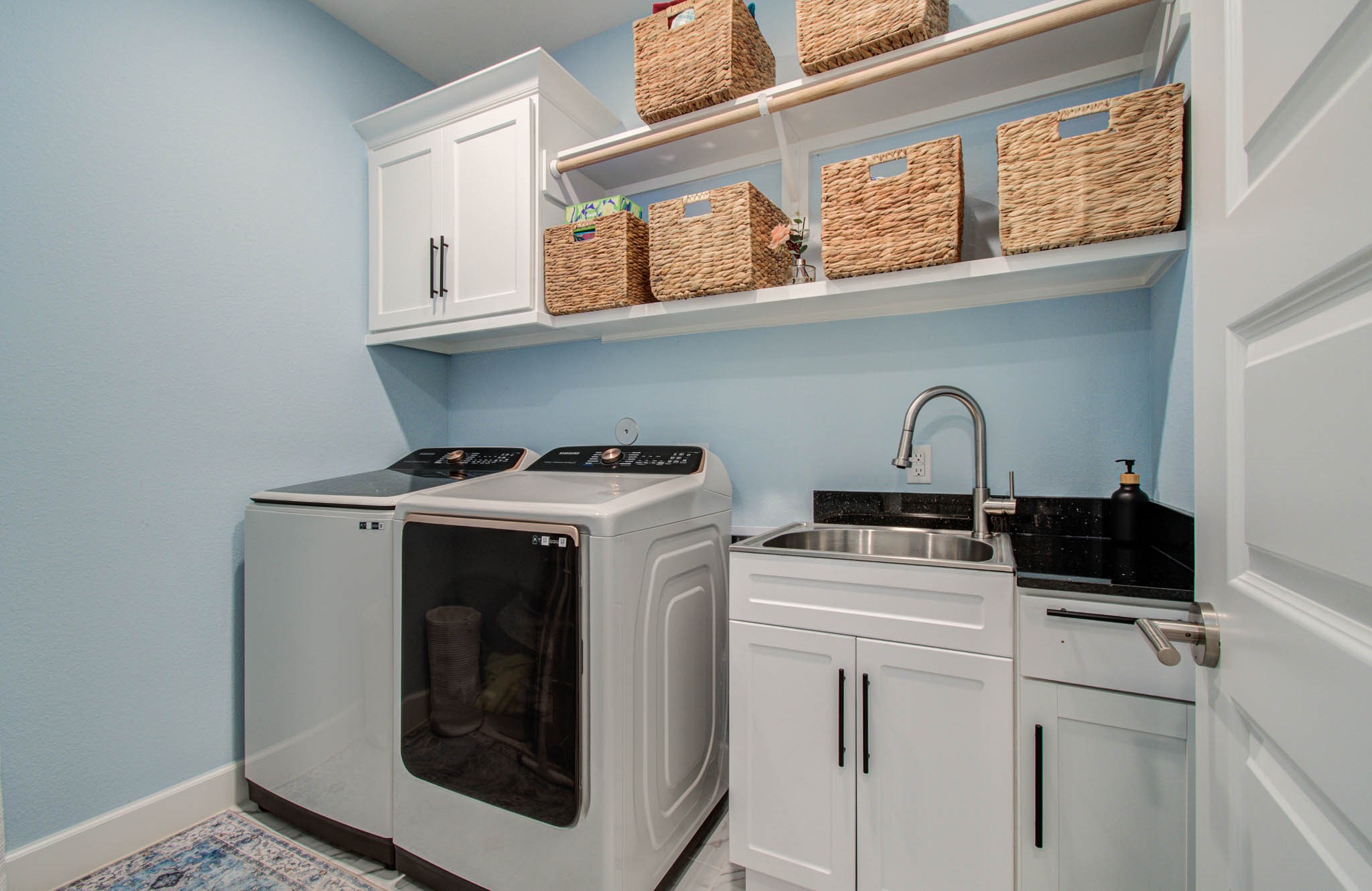 24430 Switchgrass Vly Way Katy, TX 77493 - Photo 24 of 50 This bright and functional laundry room features modern appliances, ample storage with overhead cabinets and woven baskets, a convenient sink, and a sleek black countertop, all set against a soft blue wall.