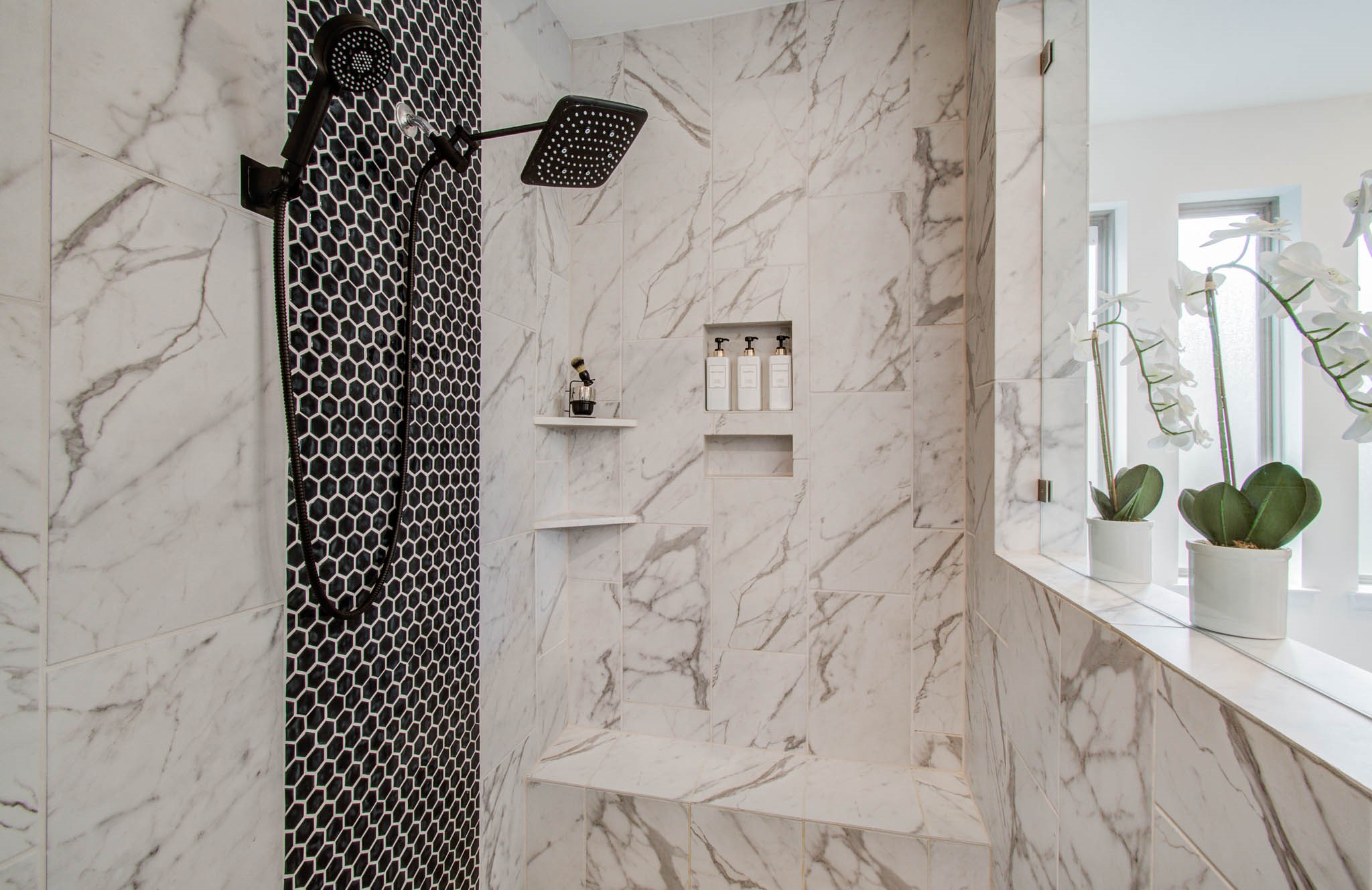 24430 Switchgrass Vly Way Katy, TX 77493 - Photo 31 of 50 This photo showcases a luxurious shower with elegant marble tiles and a striking black hexagonal accent wall. It features a modern rain showerhead, built-in shelving, and a bench for added convenience. Natural light filters in through nearby windows, enhancing the space's bright and airy feel.