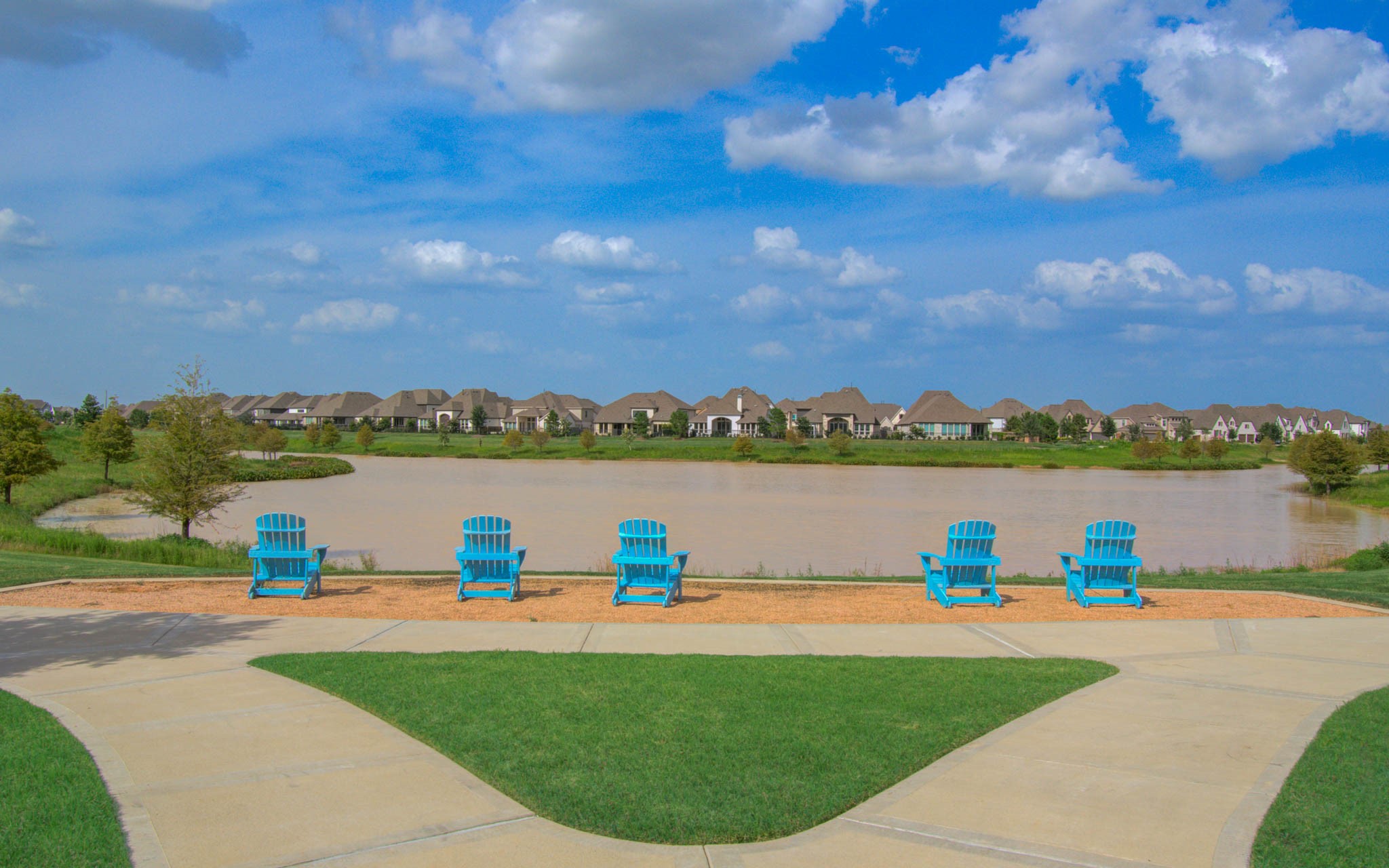 24430 Switchgrass Vly Way Katy, TX 77493 - Photo 44 of 50 A serene lakeside view with a row of inviting blue Adirondack chairs on a path, overlooking a calm water body. The backdrop features a neighborhood of picturesque homes under a bright blue sky with scattered clouds. Perfect for relaxation and enjoying the scenery.