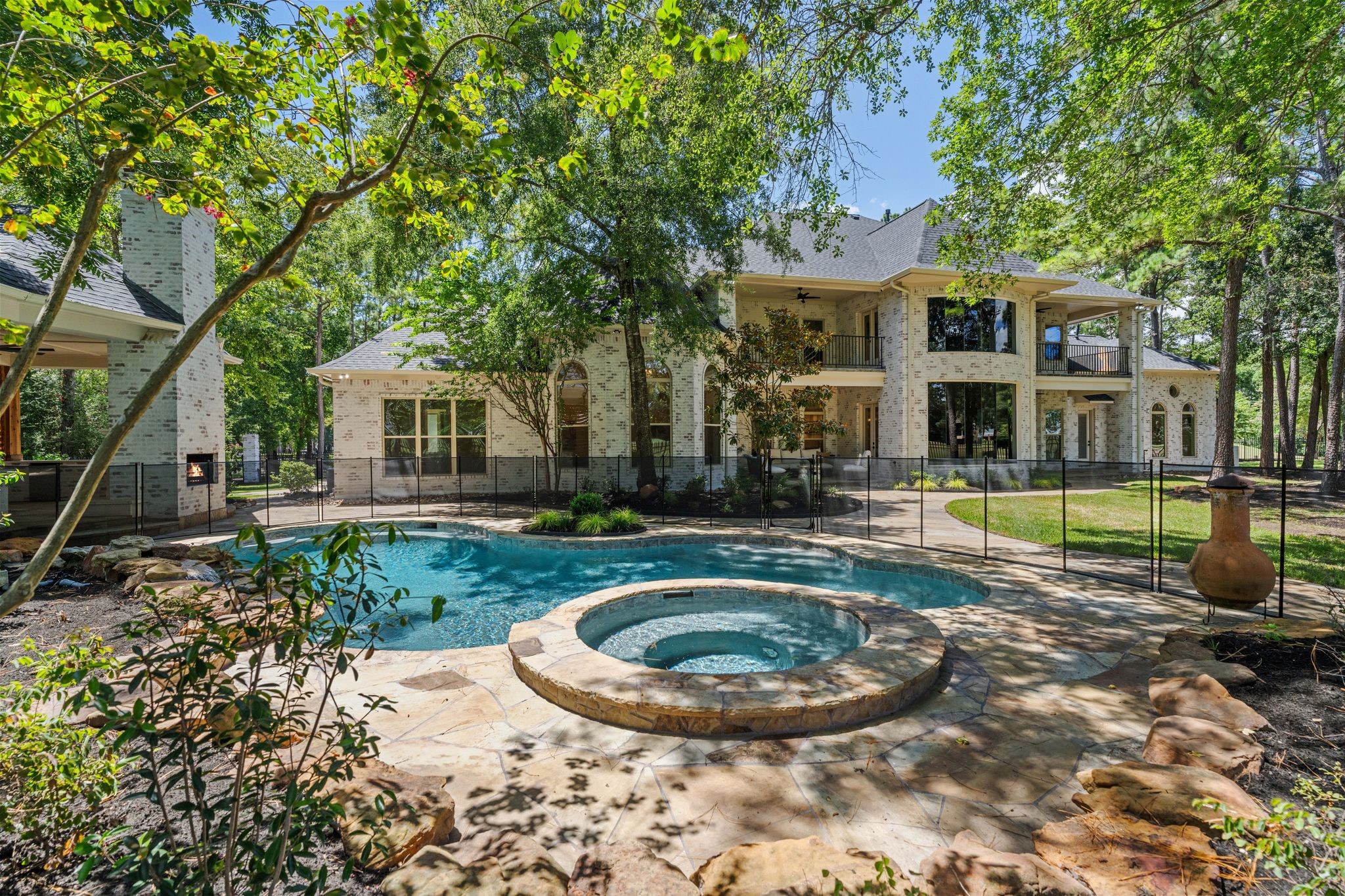 31 Hollymead Drive Spring, TX 77381 - Photo 41 of 50 This stunning home features a spacious backyard with a luxurious pool and spa, separate outdoor Pavilion with kitchen and wood burning fireplace surrounded by lush greenery and a secure removable child safety fence. The elegant brick exterior and large windows offer a perfect blend of style and comfort, ideal for relaxation and entertainment.