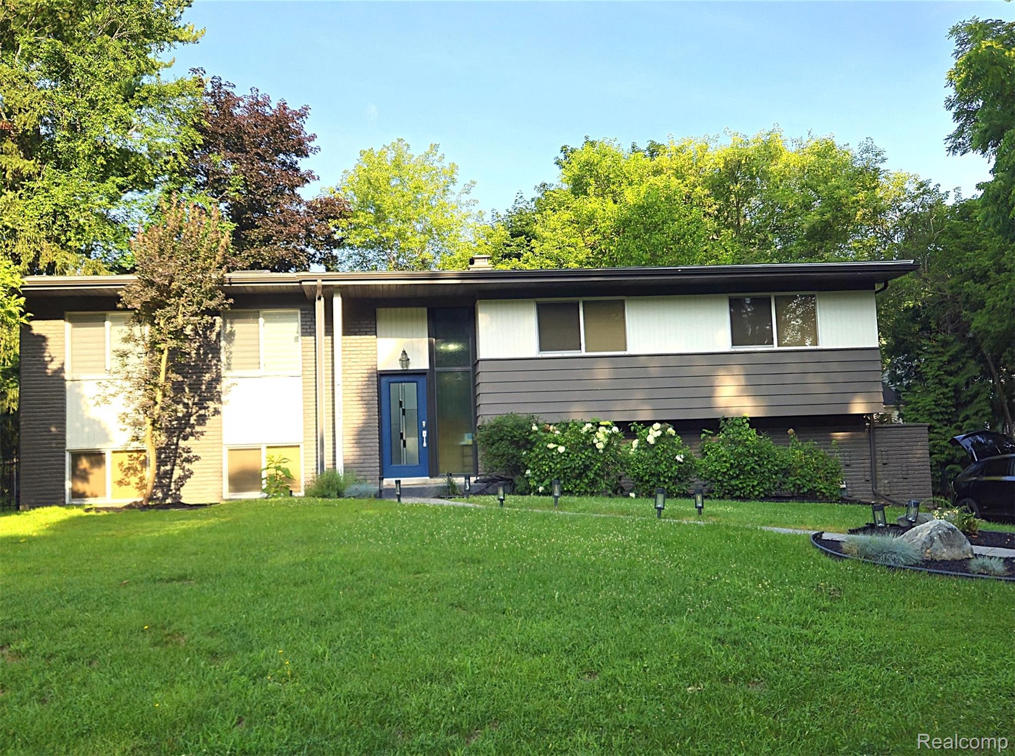 7120 Pebblecreek Road West Bloomfield, MI 48322 - Photo 2 of 31 Contemporary split-level residence featuring a blue entry door, an expansive front lawn, mature trees, and established foundational plantings
