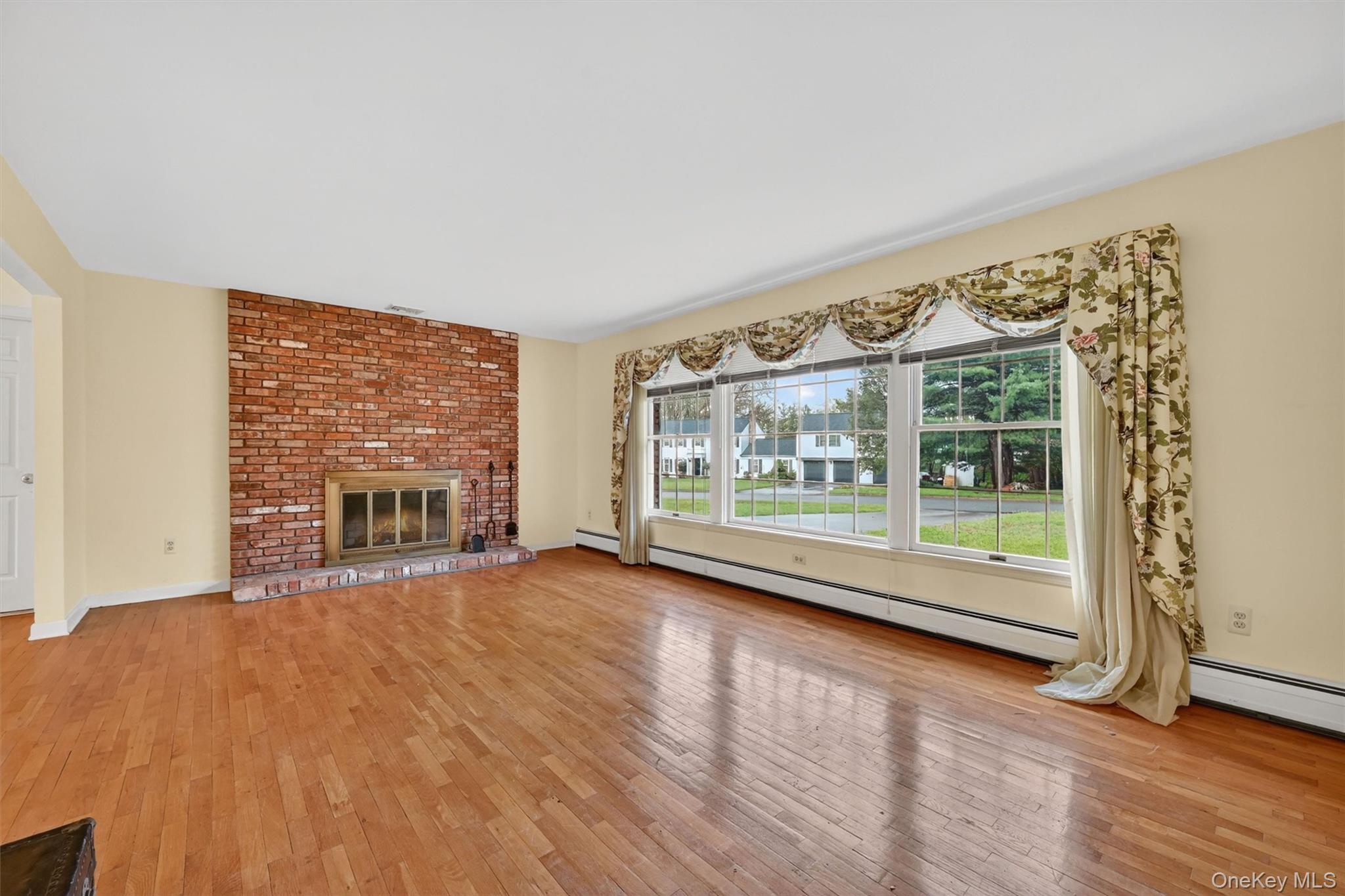 16 Ridgeview Terrace Goshen, NY 10924 - Photo 7 of 35 Spacious living area featuring a brick fireplace with a brass firebox, wood-finish flooring, and a bank of three large windows providing ample natural light