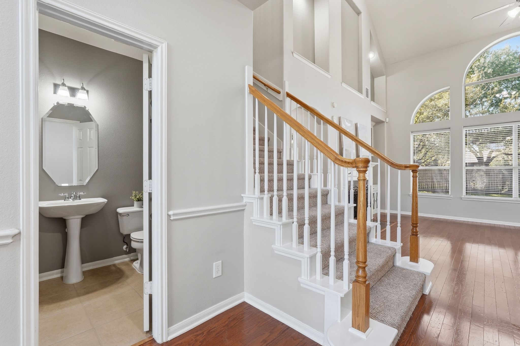 24931 Corbin Gate Drive Spring, TX 77389 - Photo 11 of 50 This photo showcases an inviting entryway with a carpeted staircase featuring wooden handrails, leading to a bright, open living area with large arched windows. Adjacent is a conveniently located powder room with a pedestal sink. The space is filled with natural light and has hardwood floors.