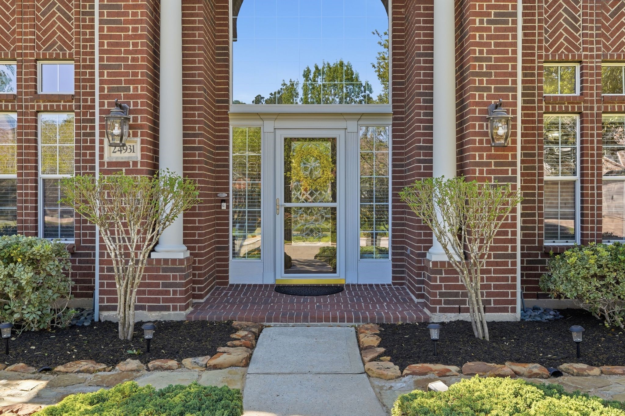 24931 Corbin Gate Drive Spring, TX 77389 - Photo 5 of 50 This photo showcases an elegant brick home entrance with large windows, white columns, and a glass front door. The landscaped front yard features well-maintained shrubs and a stone-bordered walkway.