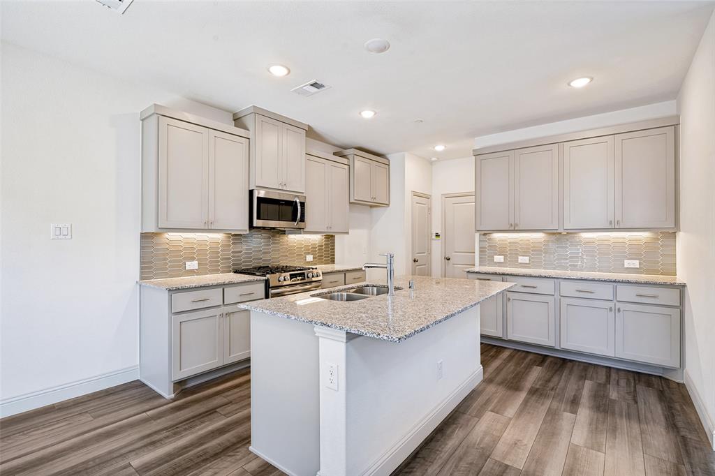1006 Emil Place Allen, TX 75013 - Photo 8 of 28 Open-concept kitchen featuring an island with a double sink, light grey cabinetry, stainless steel appliances, granite countertops, and wood-finish flooring