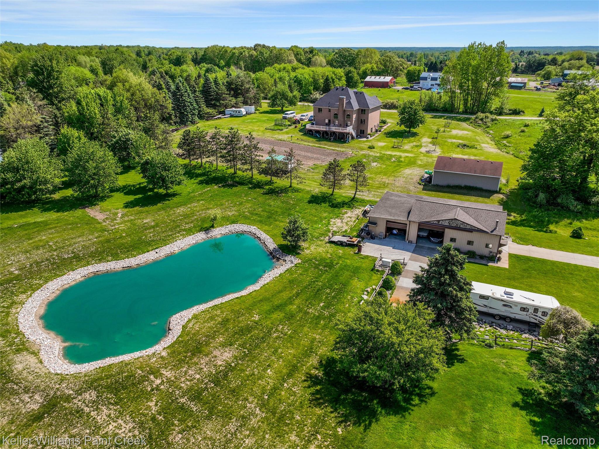 5813 36 Mile Road Romeo, MI 48065 - Photo 67 of 81 Expansive property featuring a large pond with rock-lined edges, a multi-car garage structure, a two-story residence with exterior decking, mature trees, and a separate outbuilding