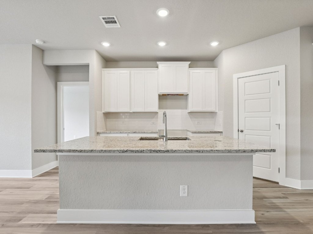 305 Gristmill Road Burnet, TX 78611 - Photo 4 of 21 Photo is not of the actual home, but is an inspirational photo of builder’s model home and may depict options, furnishings, and/or decorator features that are not includedKitchen with light stone countertops, white cabinetry, a center island with sink, light wood-style floors, and decorative backsplash