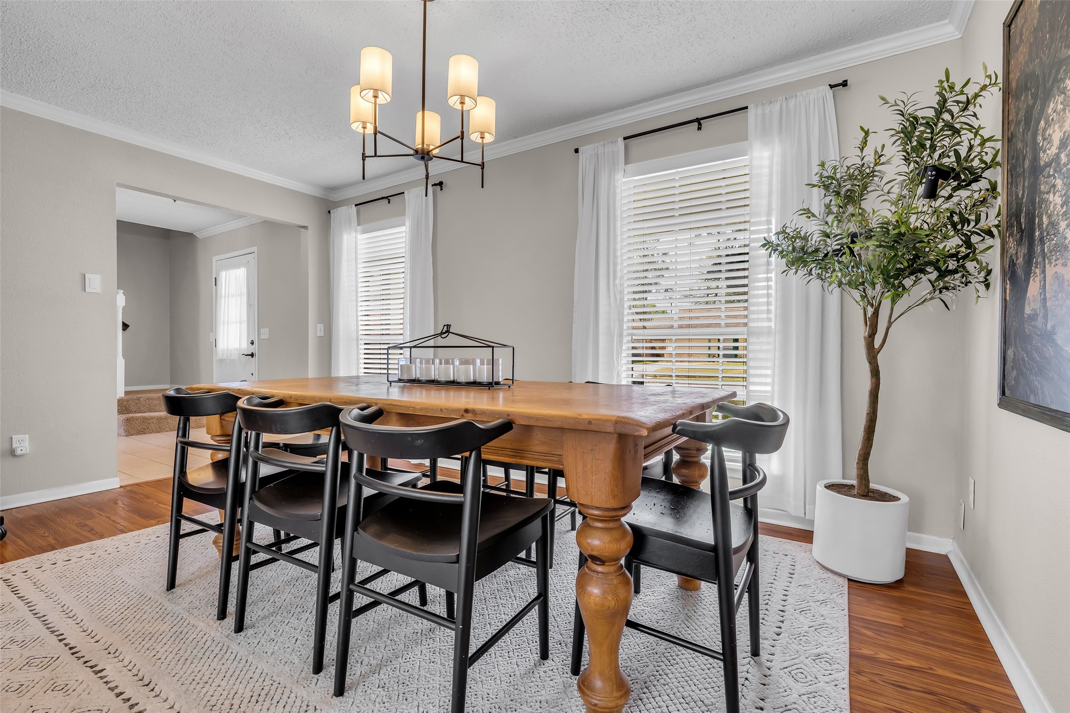 9415 Towerstone Drive Spring, TX 77379 - Photo 11 of 41 This photo showcases a well-lit dining area with a wooden table and chairs, elegant light fixture, and large artwork on the wall. The room has hardwood floors, a light neutral color palette, and large windows with white curtains, offering a bright and welcoming atmosphere.