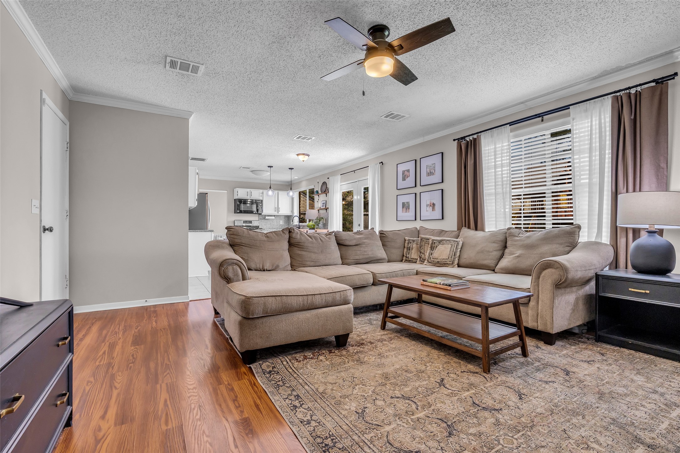 9415 Towerstone Drive Spring, TX 77379 - Photo 21 of 41 Spacious living area with a large sectional sofa, wall-mounted TV, and ceiling fan. The open layout flows into the modern kitchen, featuring white cabinetry and stainless steel appliances. Warm wood flooring and neutral tones create a cozy atmosphere.