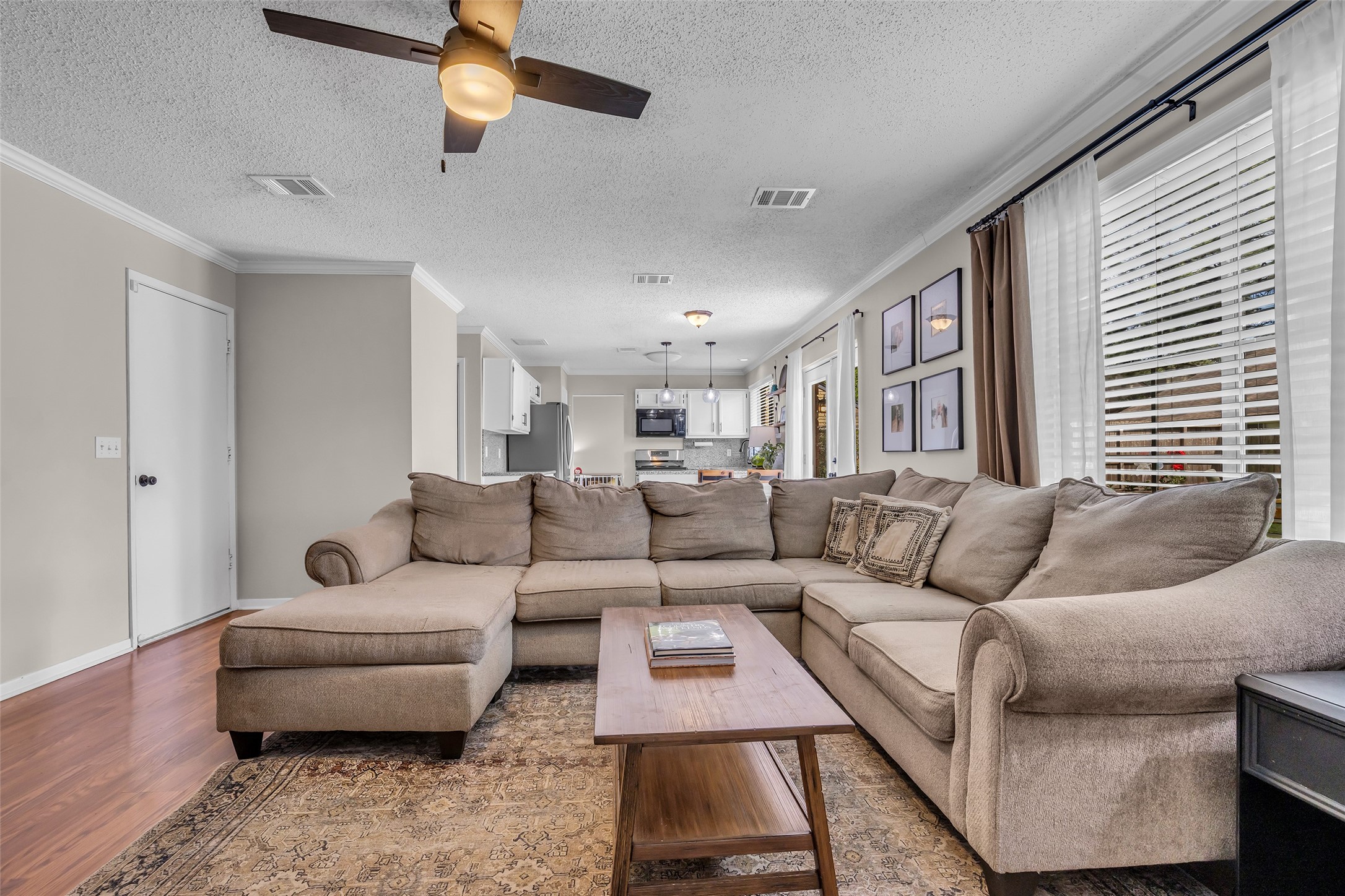 9415 Towerstone Drive Spring, TX 77379 - Photo 22 of 41 This cozy living room features a comfortable sectional sofa, a modern ceiling fan, and a large TV. The room is anchored by a white brick fireplace with a simple mantel, adorned with decorative items. Natural light streams through the large windows with elegant curtains, highlighting the warm hardwood floors.
