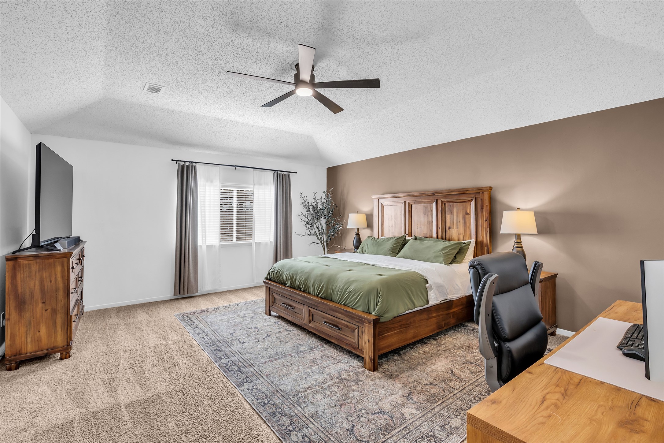 9415 Towerstone Drive Spring, TX 77379 - Photo 5 of 41 This spacious bedroom features a vaulted ceiling with a modern fan, a large window with curtains, and cozy carpeting. It includes a wooden bed with green bedding, matching nightstands with lamps, a dresser with a TV, and a desk with a chair, offering a comfortable blend of style and functionality.