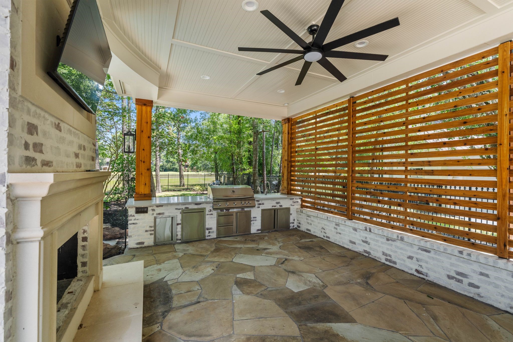 31 Hollymead Drive Spring, TX 77381 - Photo 44 of 50 This image showcases a stylish outdoor living space with a built-in barbecue, stone flooring, and a ceiling fan. The area features a modern slatted wood privacy screen and a cozy fireplace, perfect for outdoor entertaining.