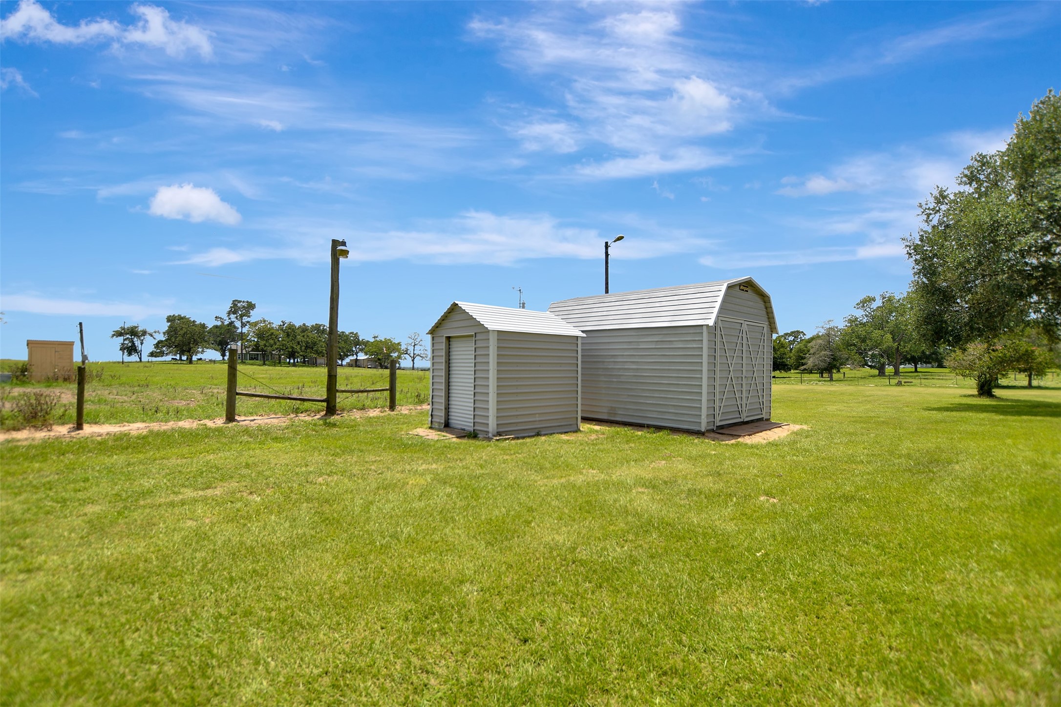 341 South S Loop Bellville, TX 77418 - Photo 32 of 33 This photo shows a spacious, grassy backyard with a small, gray storage shed and larger barn-style structure. The area is fenced and surrounded by open fields and trees, under a clear blue sky.