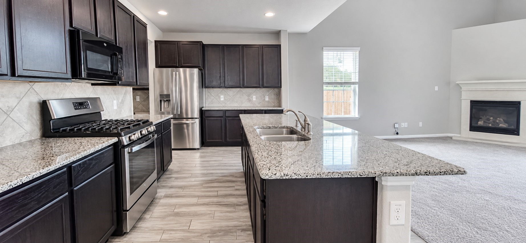 11355 Cascading Stream Way Houston, TX 77044 - Photo 6 of 17 Modern kitchen with sleek granite countertops, stainless steel appliances, and rich dark cabinetry, seamlessly connected to a cozy living area featuring a fireplace.