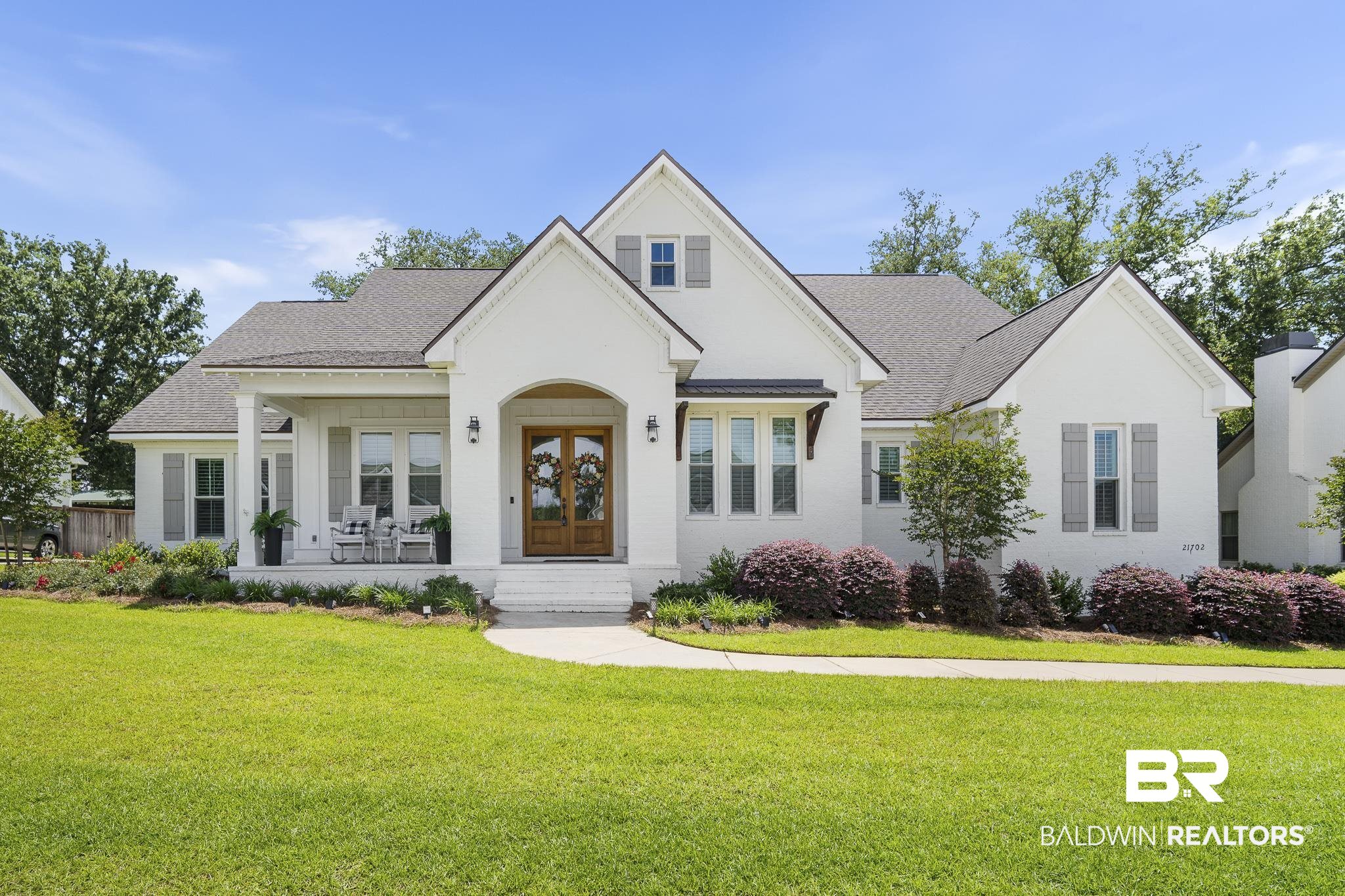 21702 Pillars Street Fairhope, AL 36532 - Photo 1 of 54 White brick exterior featuring a covered front porch, double wooden entry doors, a gabled roofline, gray window shutters, and a manicured lawn