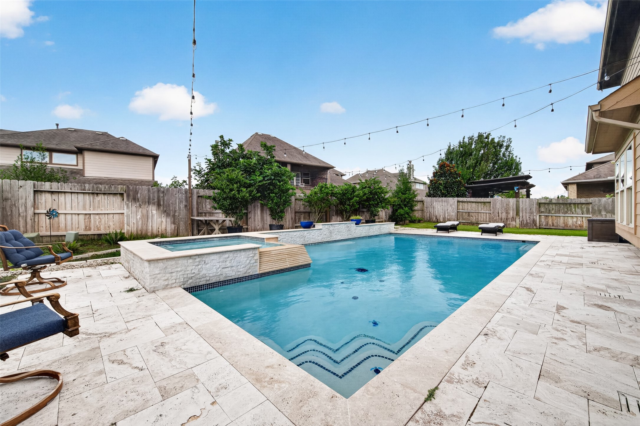 25611 Ellis Ridge Court Katy, TX 77494 - Photo 46 of 49 This photo showcases a spacious backyard with a modern swimming pool and hot tub, surrounded by a stone patio. String lights add a cozy ambiance, and there are lounge chairs for relaxation. The area is enclosed by a wooden fence, providing privacy.