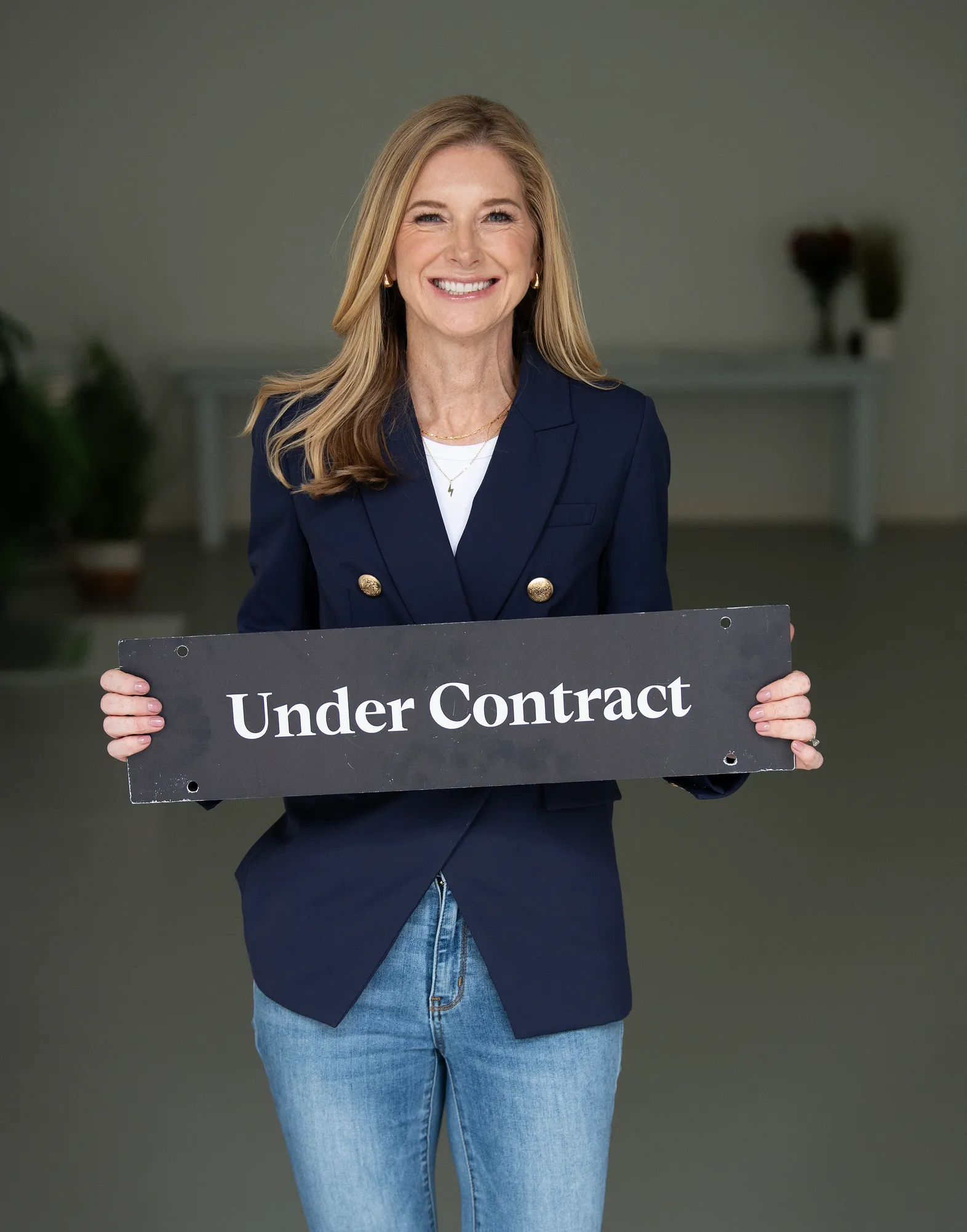 Jen Holden smiling in a navy blazer and jeans, holding an “Under Contract” sign indoors.