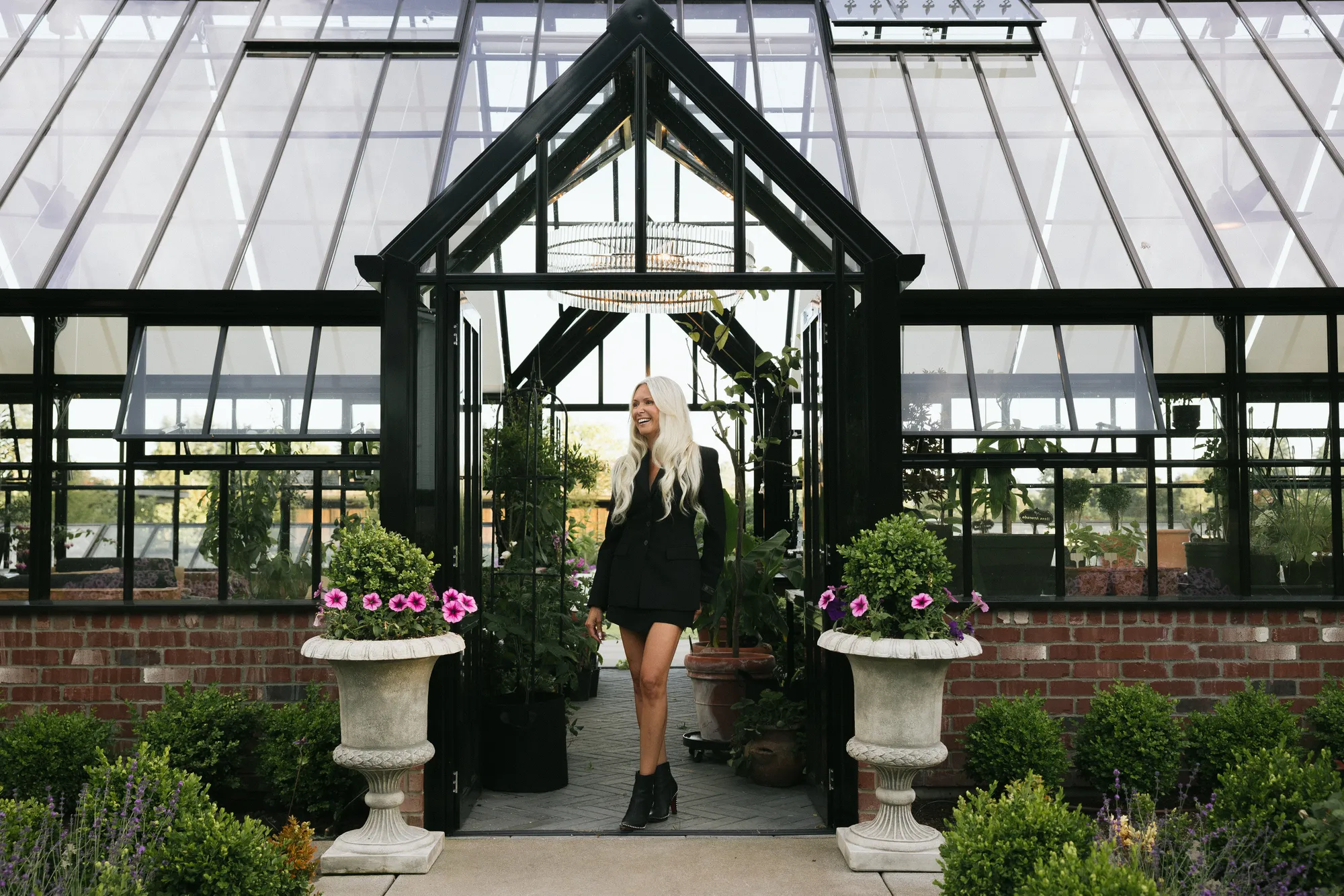 Tanya Mock, luxury real estate broker at Compass Snohomish, stands at the entrance of an elegant glass greenhouse surrounded by curated gardens, showcasing sophisticated lifestyle and estate properties in Snohomish County.