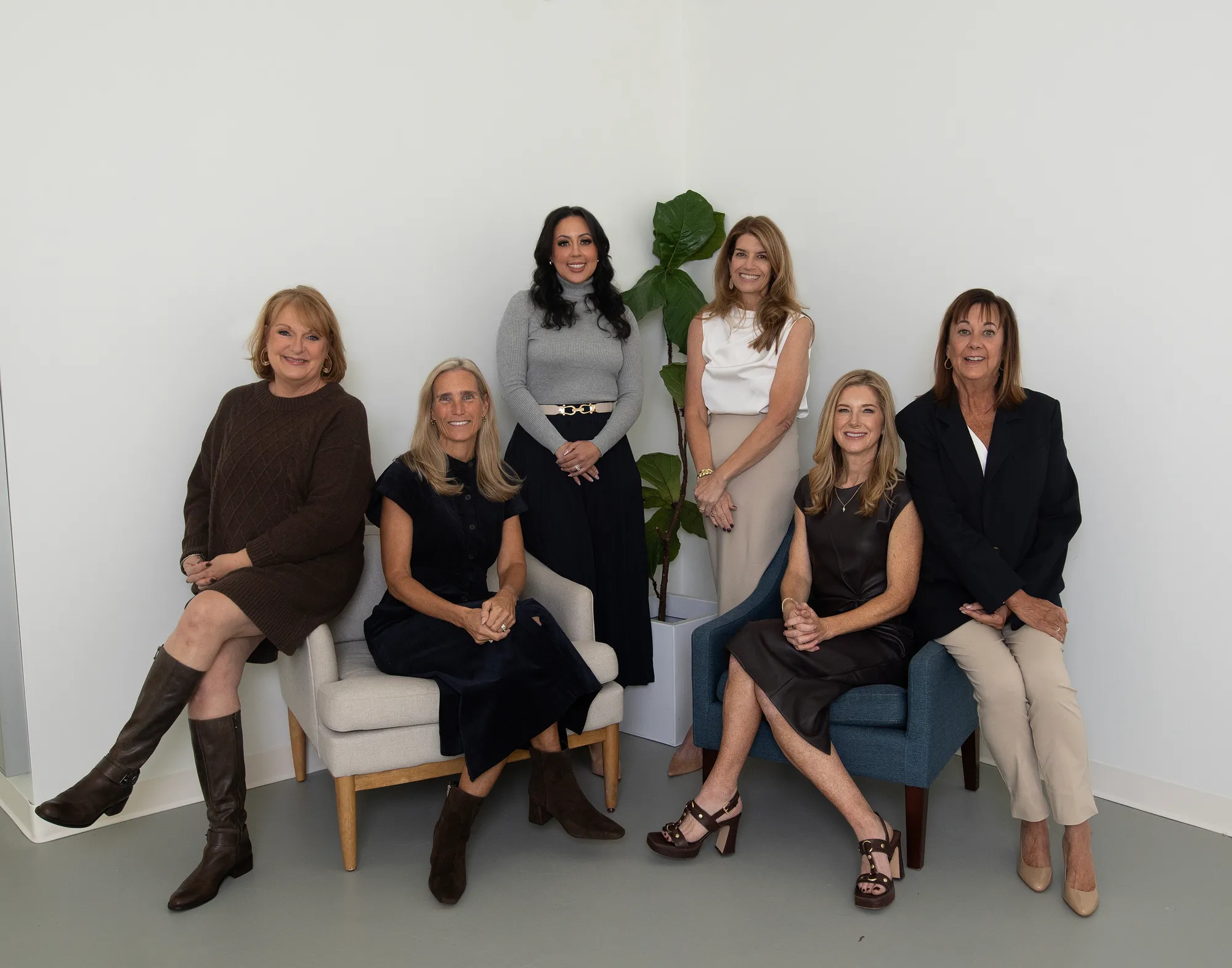 Jen Holden Group of Compass team portrait with six women in professional attire, seated and standing in a bright, modern studio with neutral chairs and a leafy plant.