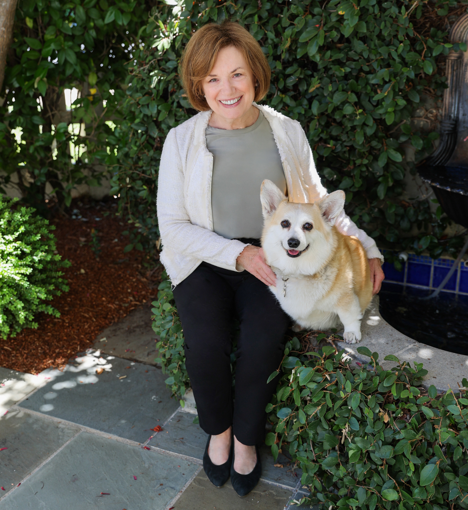 Judy sits outside in a garden setting against a background of greenery. Her beloved corgi sits next to her. Both Judy and the pup are facing the camera and smiling.
