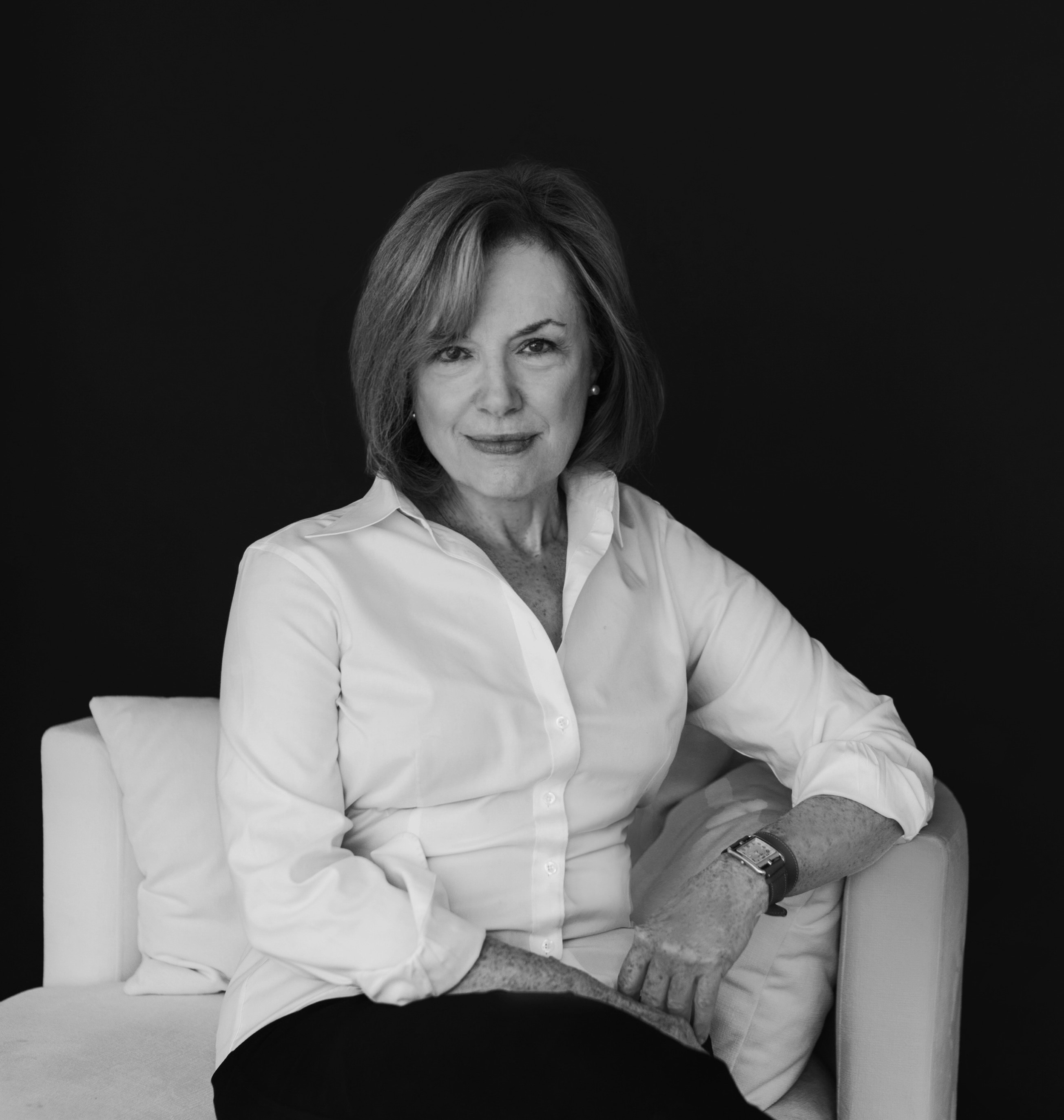 black and white portrait of Judy. She's dressed in business casual attire and sits on a plush armchair. She's looking directly into the camera, with a confident smile.