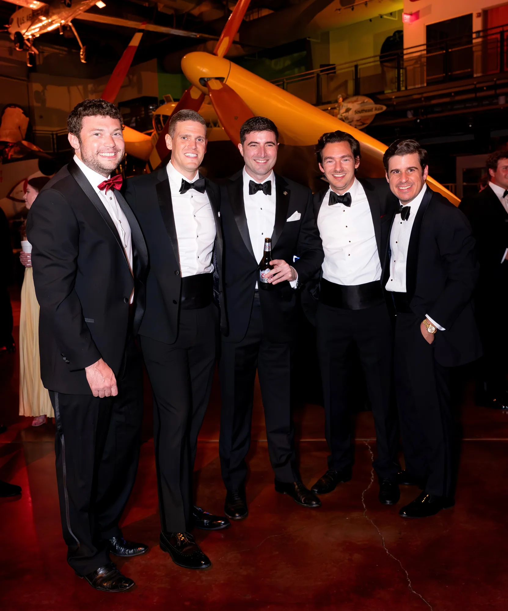 5 men wearing tuxedos in an airplane hangar