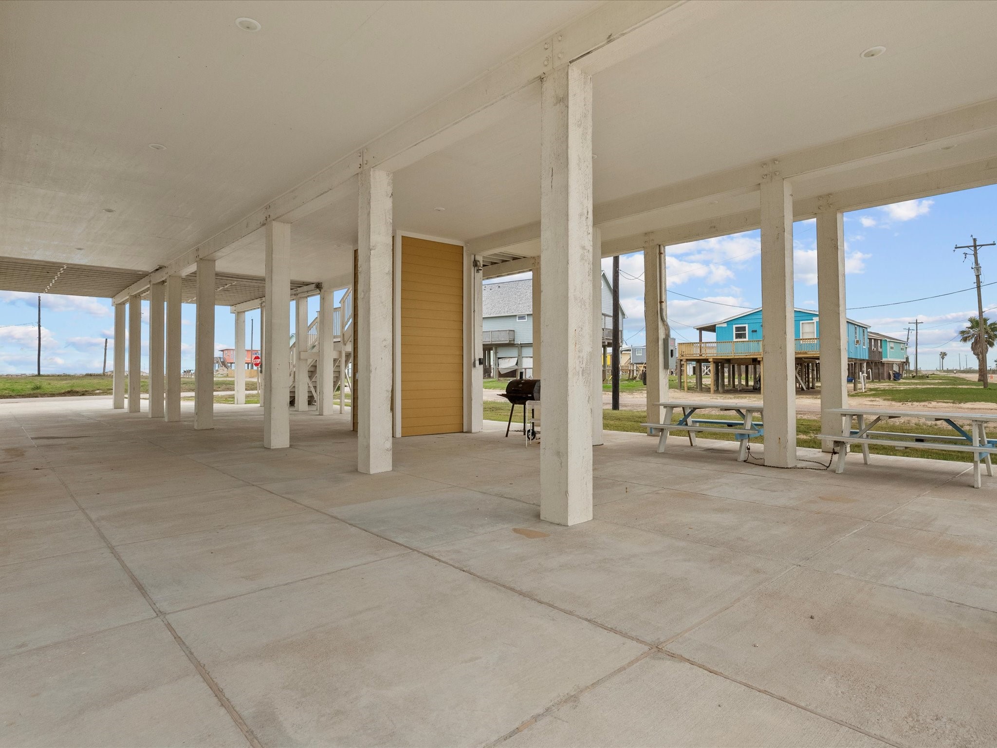 1018 Seashell Drive Surfside Beach, TX 77541 - Photo 49 of 49 This photo shows an elevated home with an open, spacious ground level featuring concrete flooring and sturdy columns. Ideal for parking or outdoor gatherings, it includes picnic tables and a grill setup. The surrounding area features stilted houses and a coastal vibe.