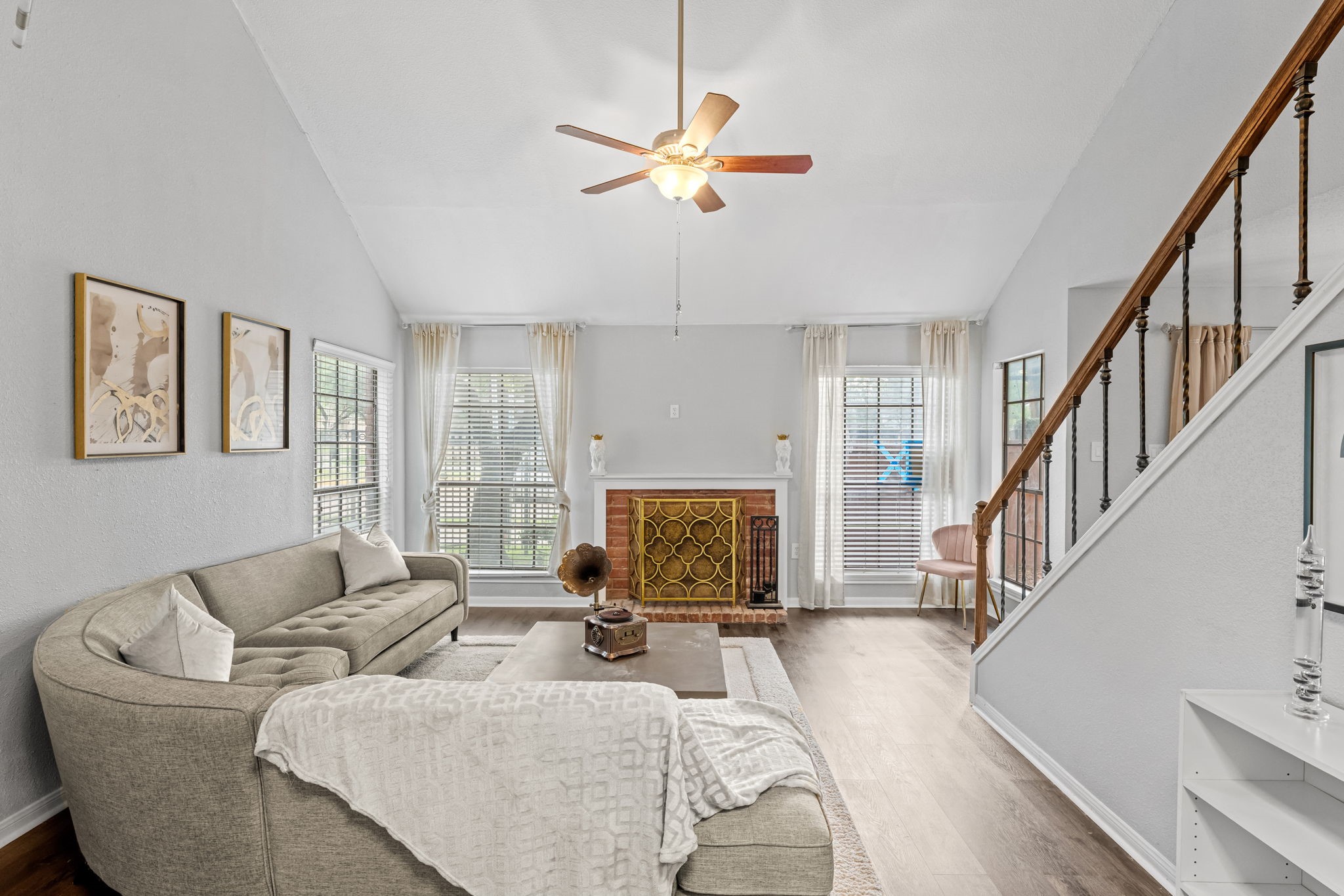 2211 South Kirkwood Road, Unit 1 Houston, TX 77077 - Photo 1 of 16 Welcome to 2211 S Kirkwood Road, Unit 1 — a two-story corner unit in the gated Lakeside Green community where the living room sets the tone immediately. Vaulted ceilings rise to a peaked apex above a wood-burning brick fireplace centered on the far wall, flanked by windows on both sides that fill the room with natural light. Dark wood spindle staircase railing frames the right edge of the space, reinforcing the townhome-style character this unit delivers from the moment you walk in.