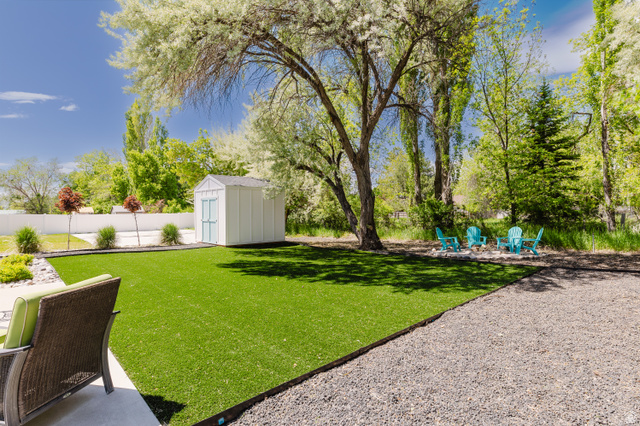 287 East 1000 North Logan, UT 84321 - Photo 36 of 41 Expansive backyard featuring artificial turf, a white privacy fence, a storage shed with light blue doors, mature trees, and a gravel patio area