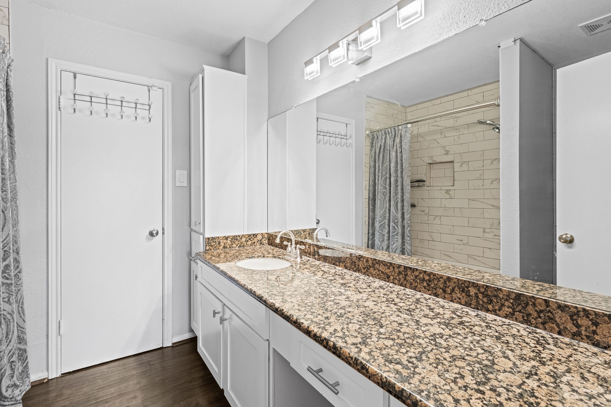 2211 South Kirkwood Road, Unit 1 Houston, TX 77077 - Photo 5 of 16 The primary bath is anchored by a wide dark speckled granite vanity that runs nearly the full width of the wall — substantially more counter surface than a standard condo bath delivers. White shaker cabinetry, a full-width mirror, and a modern rectangular light bar complete the vanity wall, with white subway tile in the shower visible in the mirror's reflection.