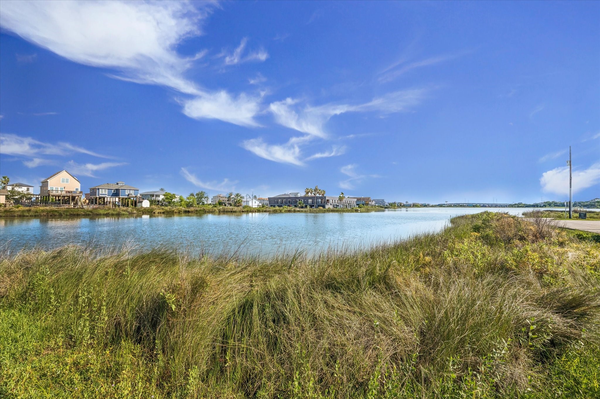 0 Hester Street Seabrook, TX 77586 - Photo 7 of 16 This photo showcases a serene waterfront view with houses along the shoreline under a clear blue sky. The foreground features lush grasses and vegetation, creating a tranquil, natural setting ideal for a peaceful lifestyle.