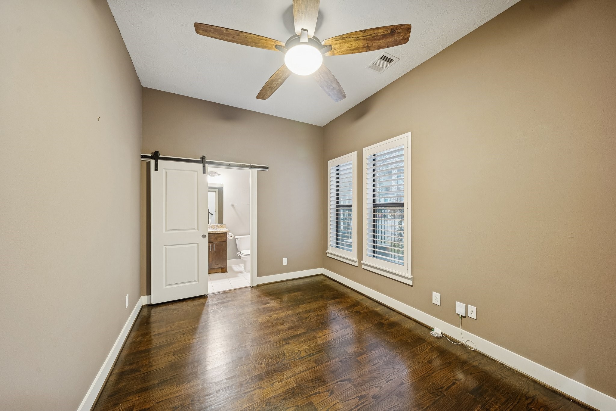 2105 Mason Street Houston, TX 77006 - Photo 20 of 43 Secondary bedroom with hardwood floors, a modern ceiling fan, and two windows with blinds that bring in plenty of natural light. A sliding barn-style door adds a stylish touch and provides convenient access to the adjacent bath, creating an easy, functional setup for guests or everyday living.