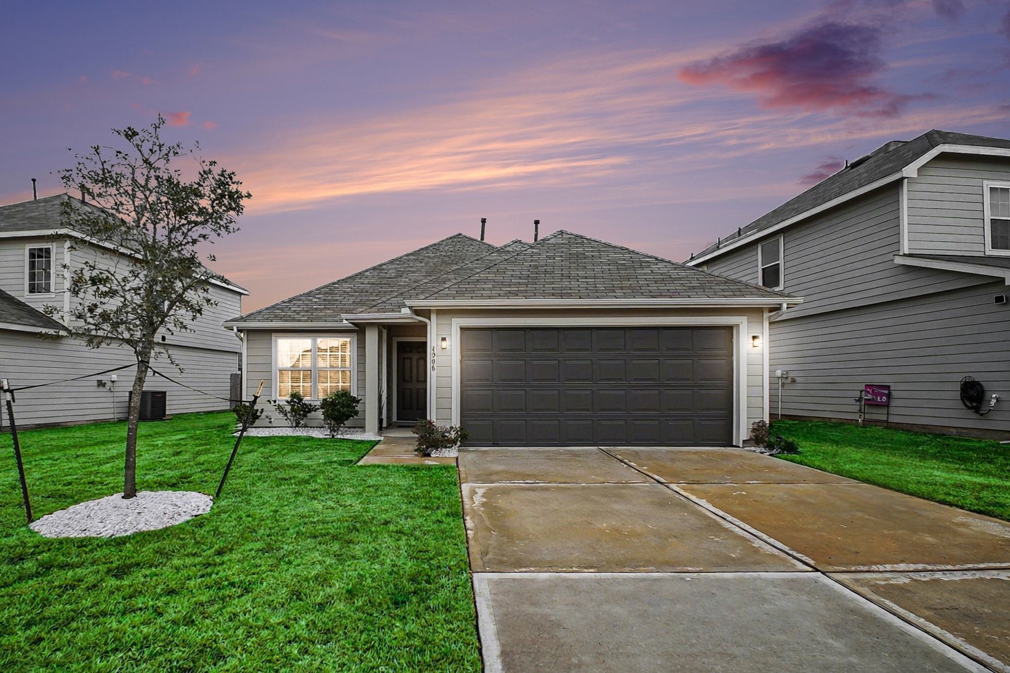 Charming single-story home with a neatly maintained front lawn with a sapling for future shade, and a spacious driveway leading to a two-car garage. The exterior is modern and inviting, set against a beautiful sunset sky.
