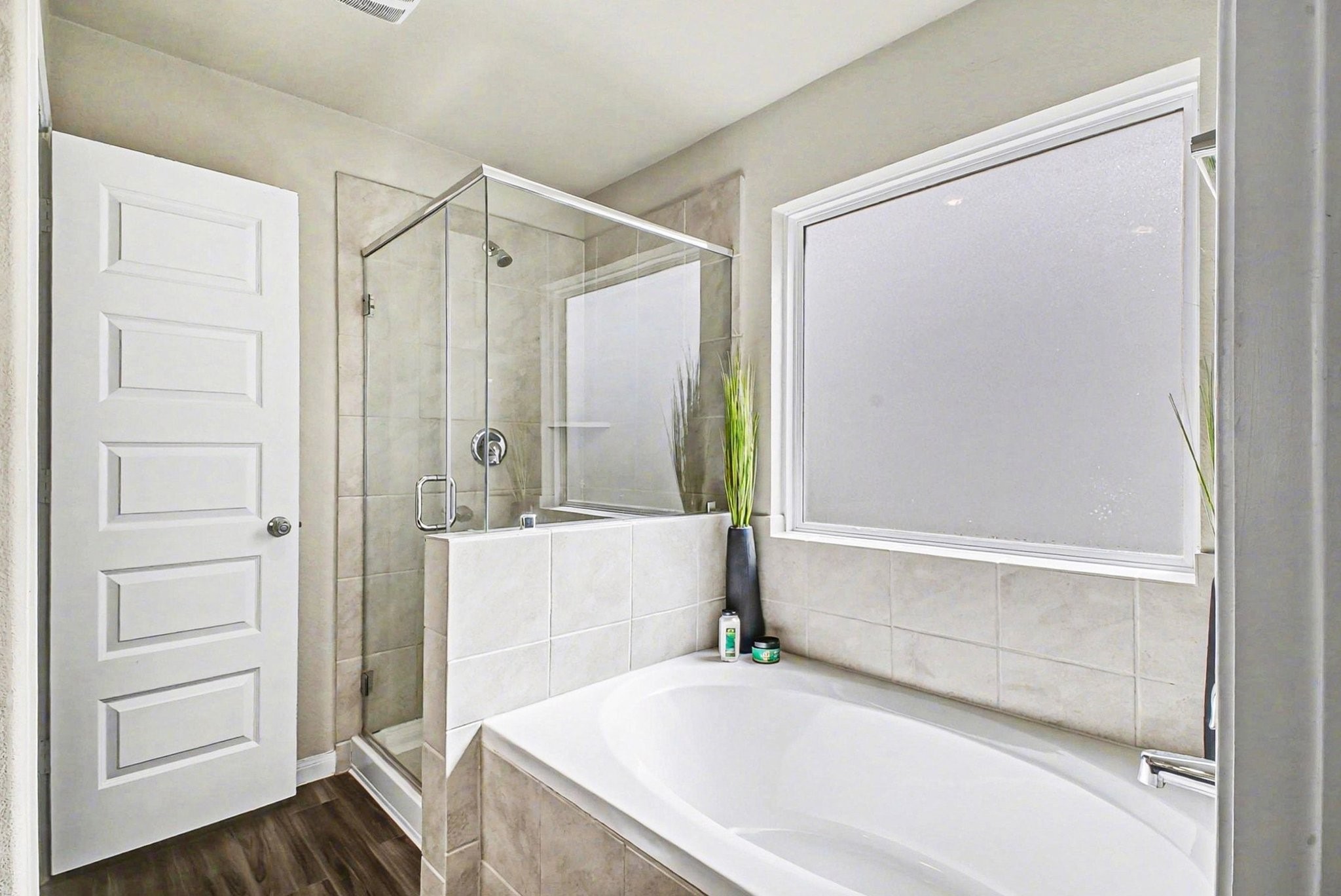 4906 Luke Matthew Drive Rosharon, TX 77583 - Photo 17 of 34 This bathroom features a modern design with a spacious bathtub and a separate glass-enclosed shower. It has a large frosted window for privacy and natural light, tiled walls, and a sleek wood-like floor.