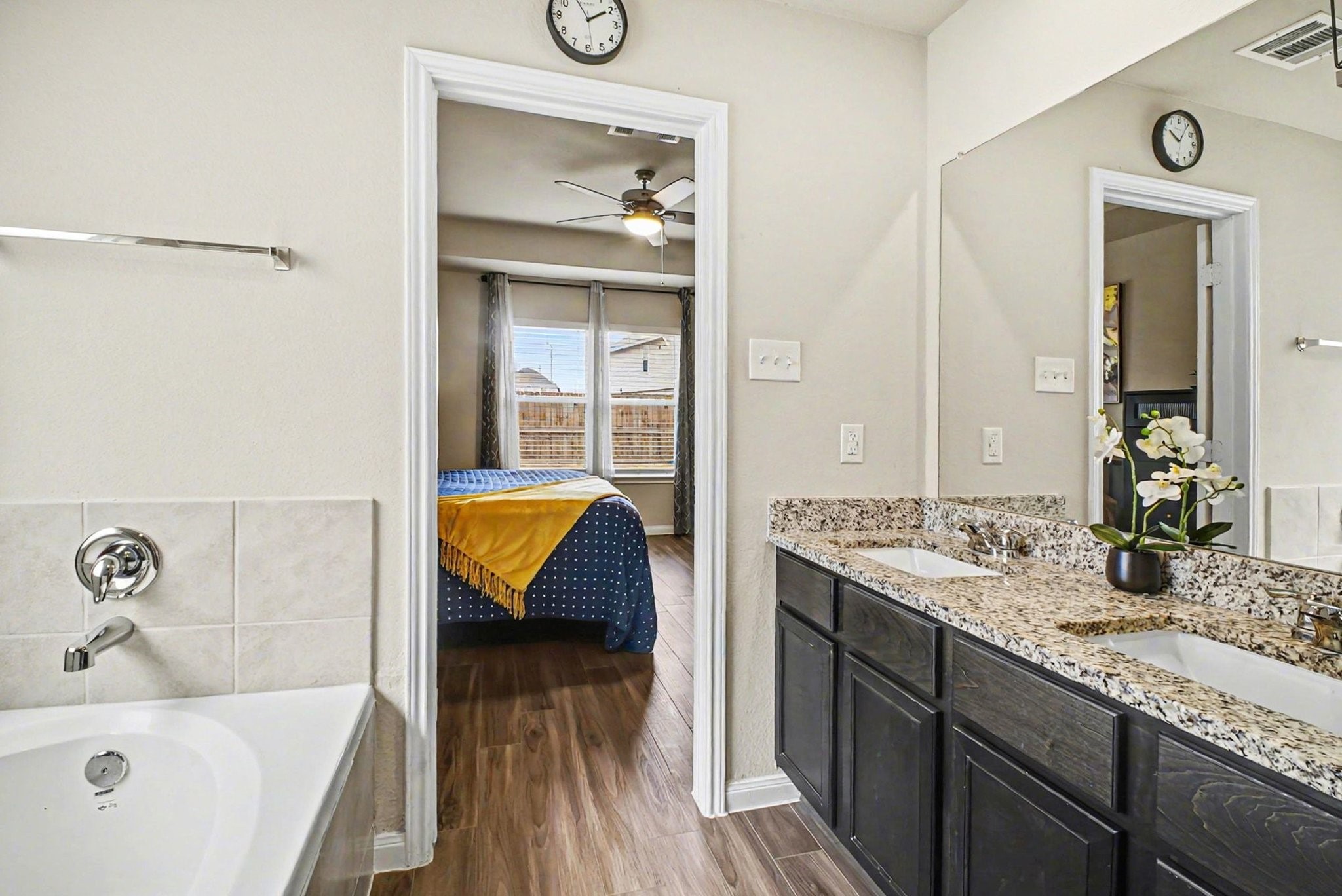 4906 Luke Matthew Drive Rosharon, TX 77583 - Photo 19 of 34 Primary modern bathroom with a granite double vanity, dark cabinets, and a spacious bathtub. It connects to a bright bedroom featuring wood flooring, large windows, and a ceiling fan.