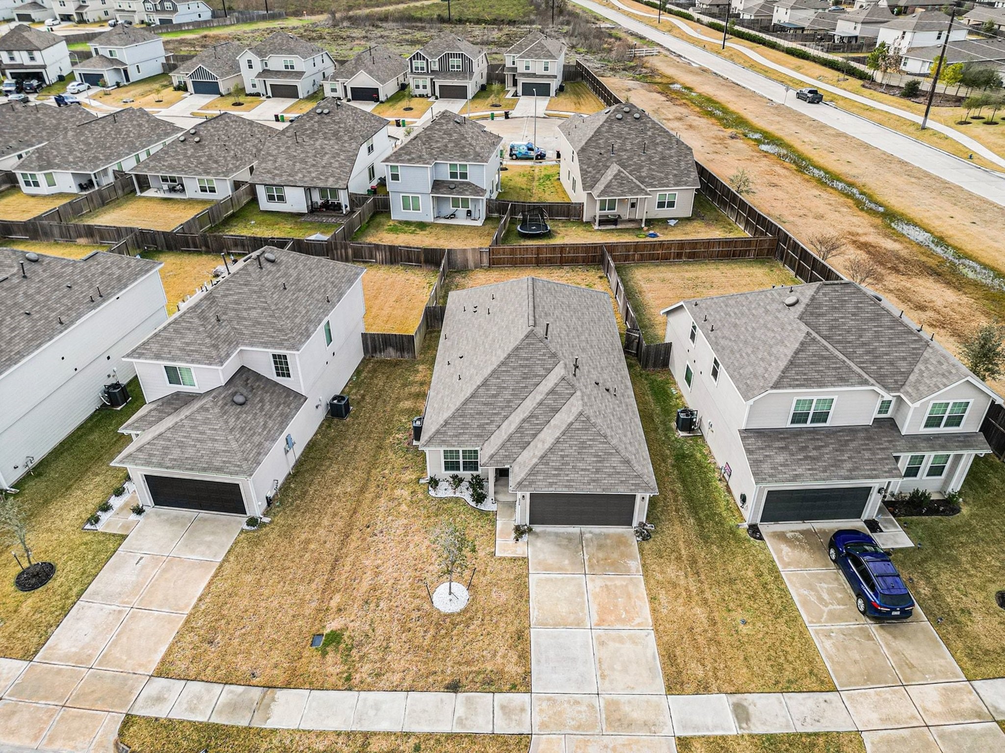 4906 Luke Matthew Drive Rosharon, TX 77583 - Photo 30 of 34 This aerial view shows a suburban neighborhood with modern two-story homes featuring gray roofs and driveways. The houses are spaced evenly with fenced backyards, and a main road runs nearby. The area appears well-maintained and residential.