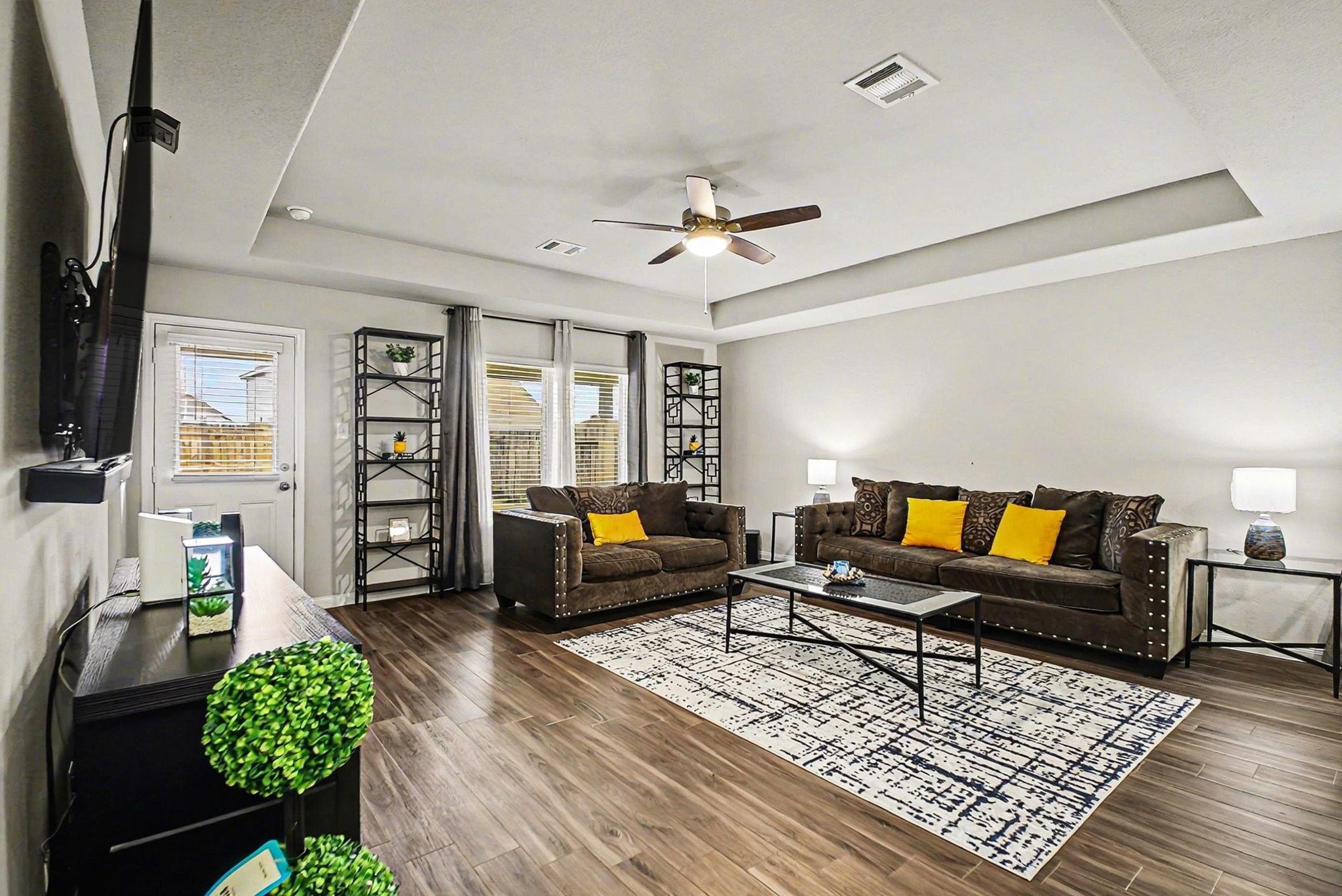 4906 Luke Matthew Drive Rosharon, TX 77583 - Photo 3 of 34 This spacious living room features stylish wood flooring, a tray ceiling with a modern fan, and ample natural light from large windows. It includes comfortable seating, a wall-mounted TV, and tasteful decor, creating a welcoming and contemporary atmosphere.