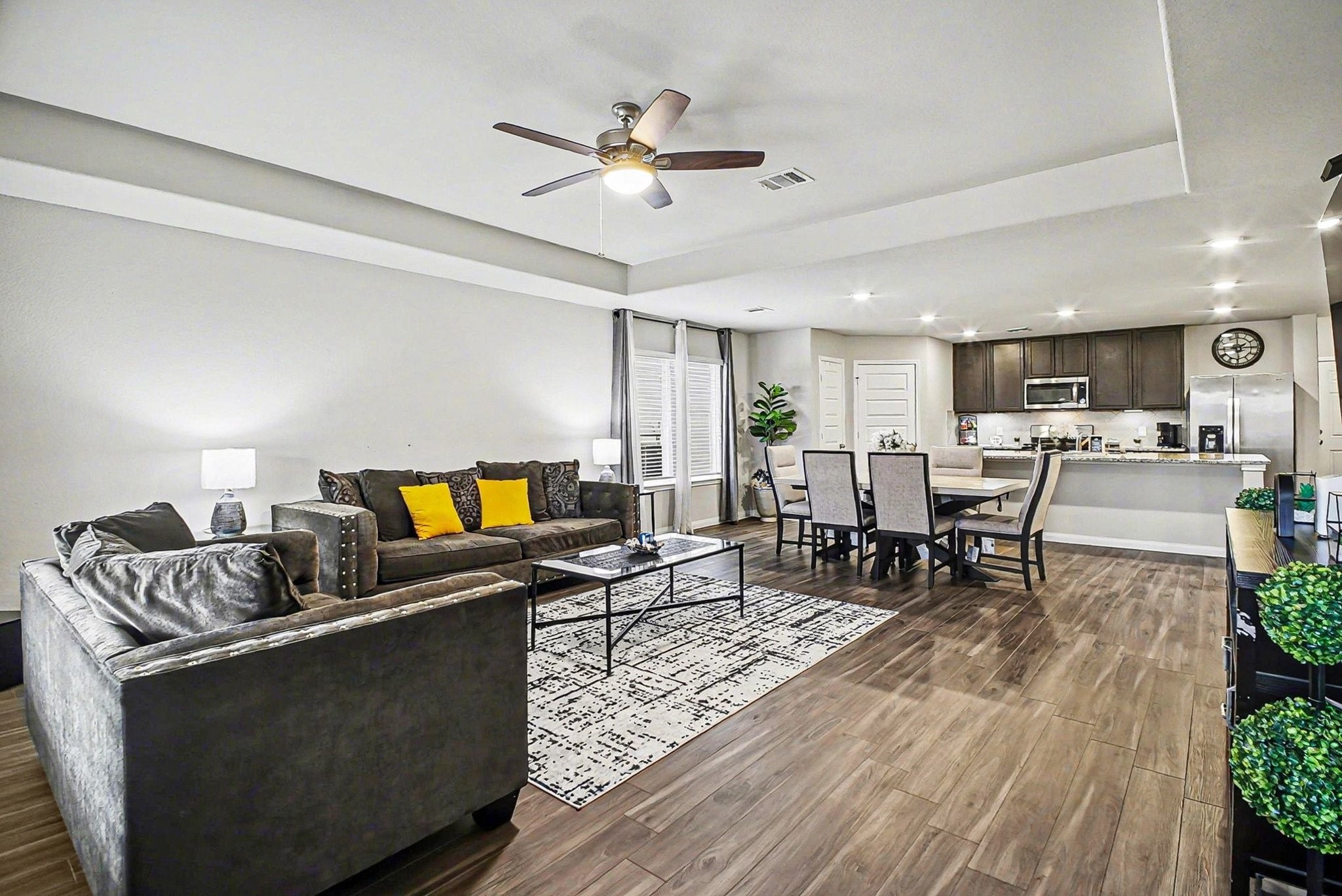 4906 Luke Matthew Drive Rosharon, TX 77583 - Photo 4 of 34 The photo showcases a spacious open-concept living area with modern decor. It features a comfortable seating arrangement with plush sofas, a dining area with a table and chairs, and a contemporary kitchen with stainless steel appliances and dark cabinetry. The room is well-lit with natural light and recessed lighting, and the wood-look flooring adds warmth.
