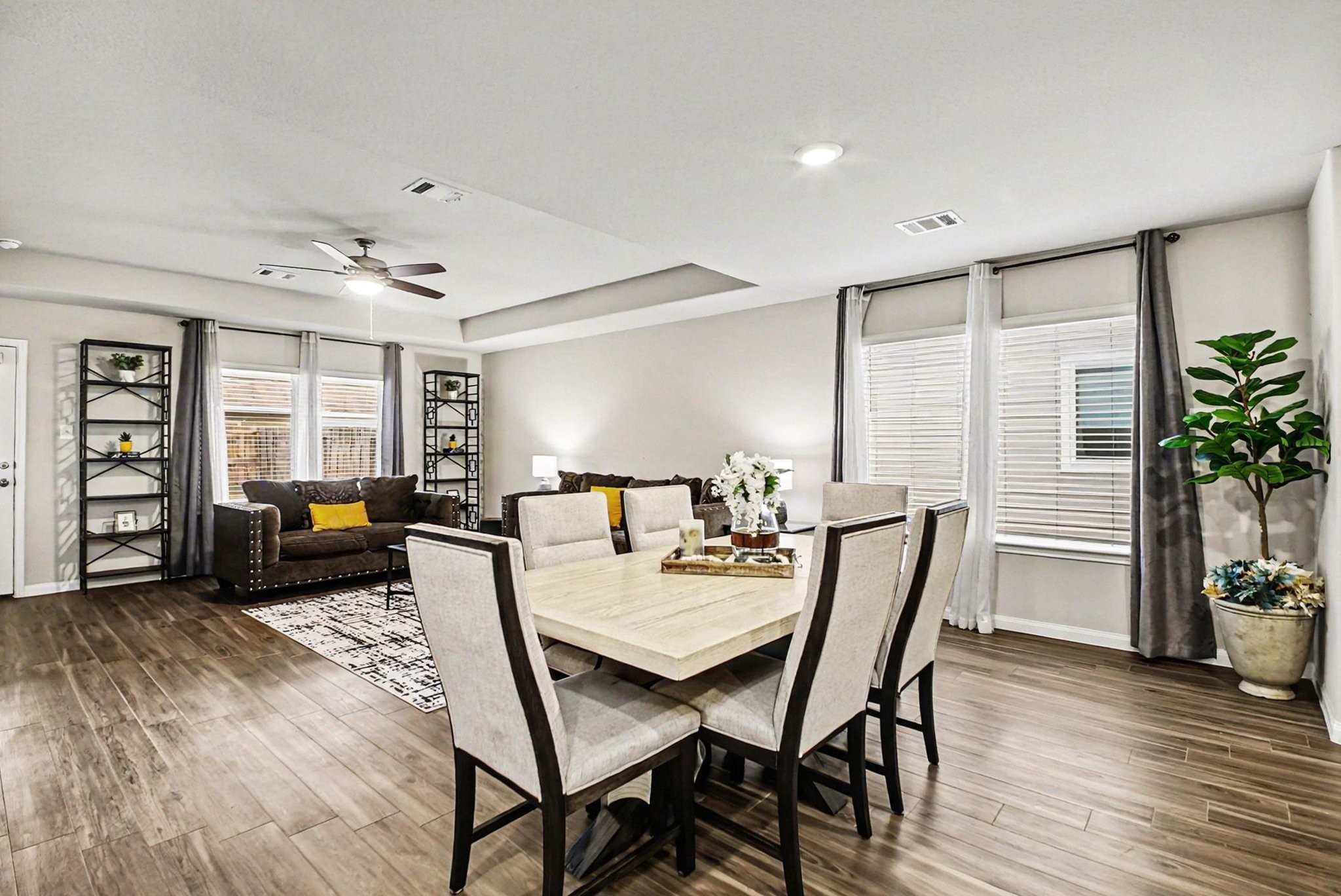 4906 Luke Matthew Drive Rosharon, TX 77583 - Photo 5 of 34 This spacious open-concept living and dining area features wood-style flooring, neutral tones, and ample natural light. The room is tastefully furnished with a cozy seating area, modern dining set, and decorative shelving. Large windows with curtains and a ceiling fan enhance the inviting atmosphere.